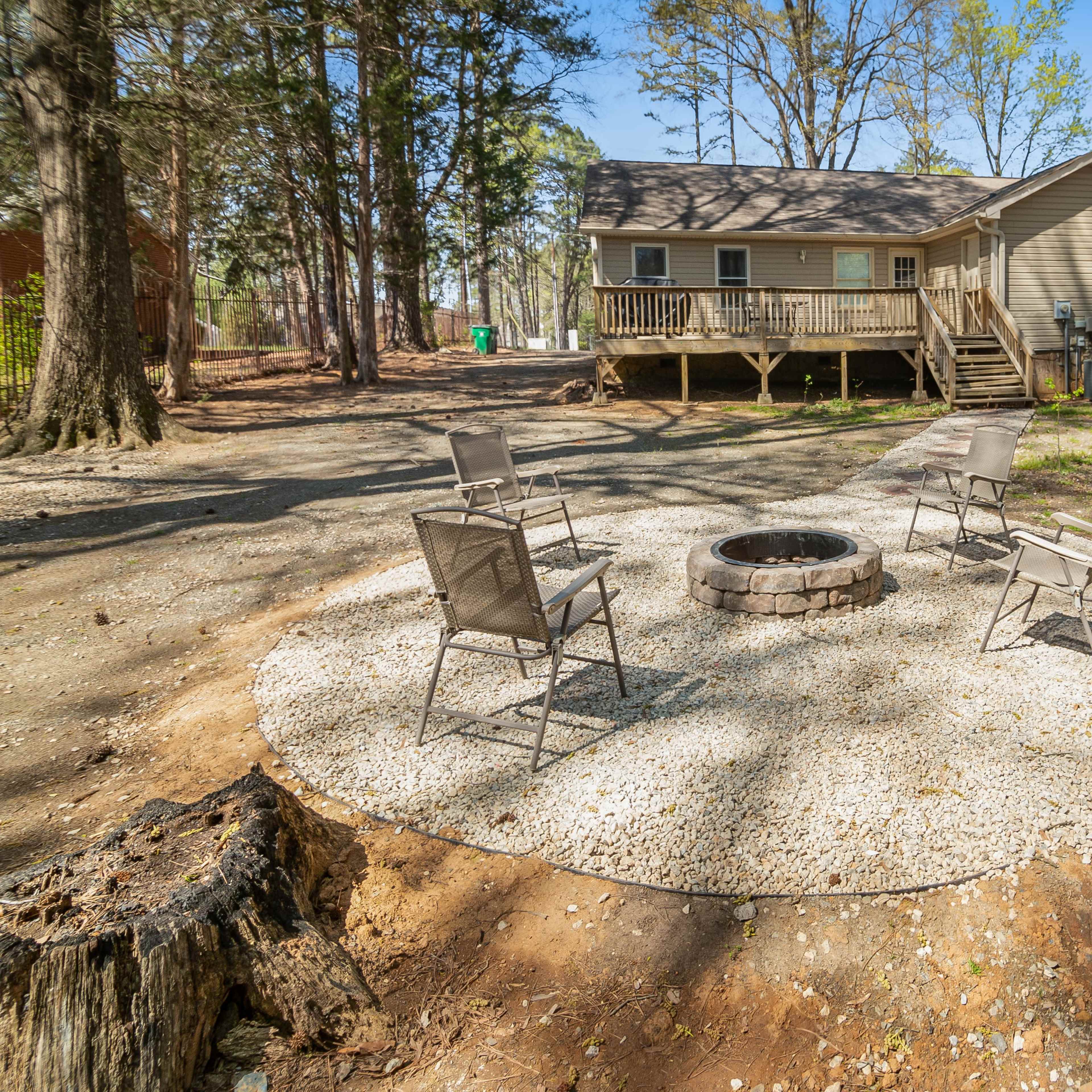 A circular gravel patio with four chairs surrounds a fire pit, located in a wooded backyard beside a house.