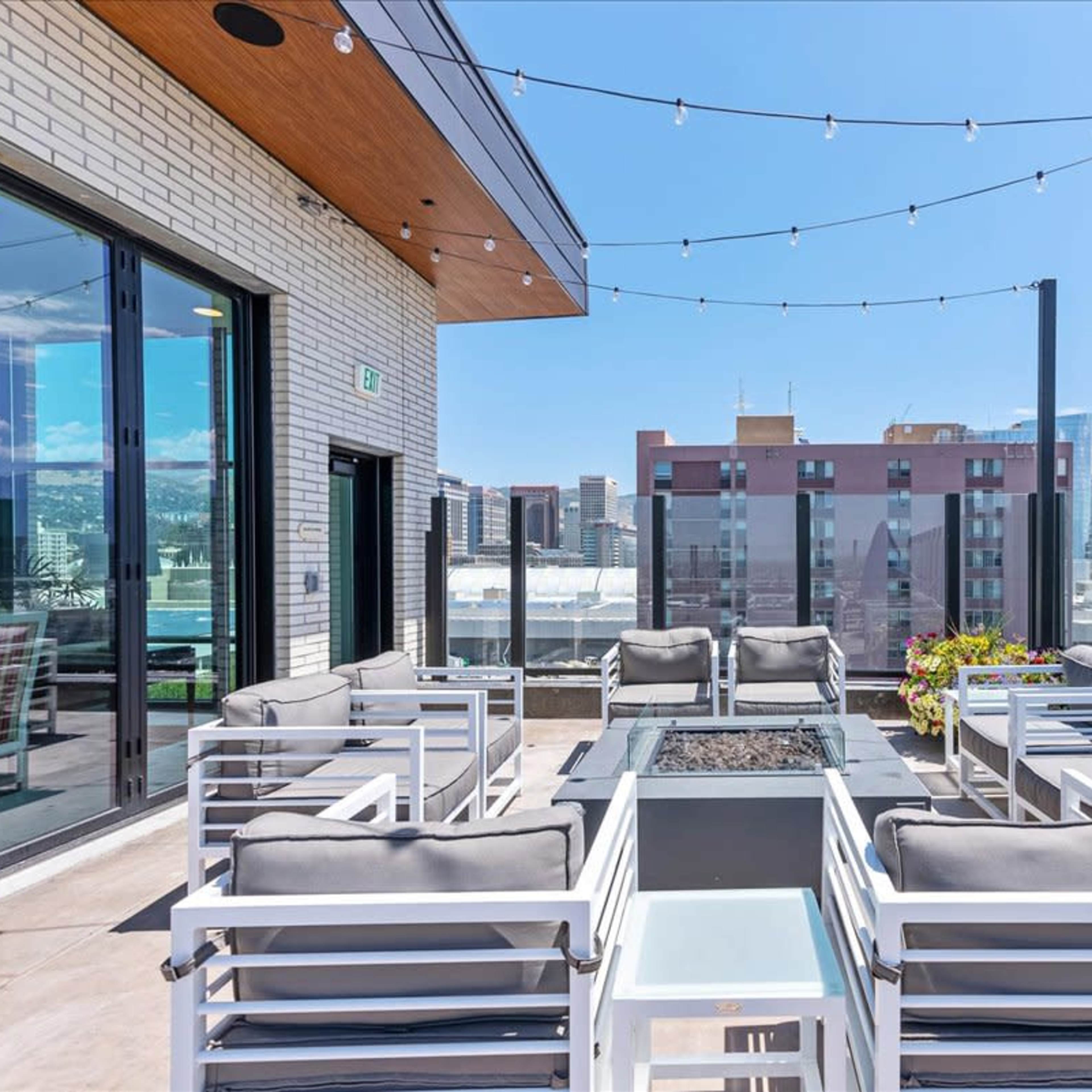 A rooftop terrace features a fire pit surrounded by white outdoor furniture and string lights overhead, with a city skyline in the background.