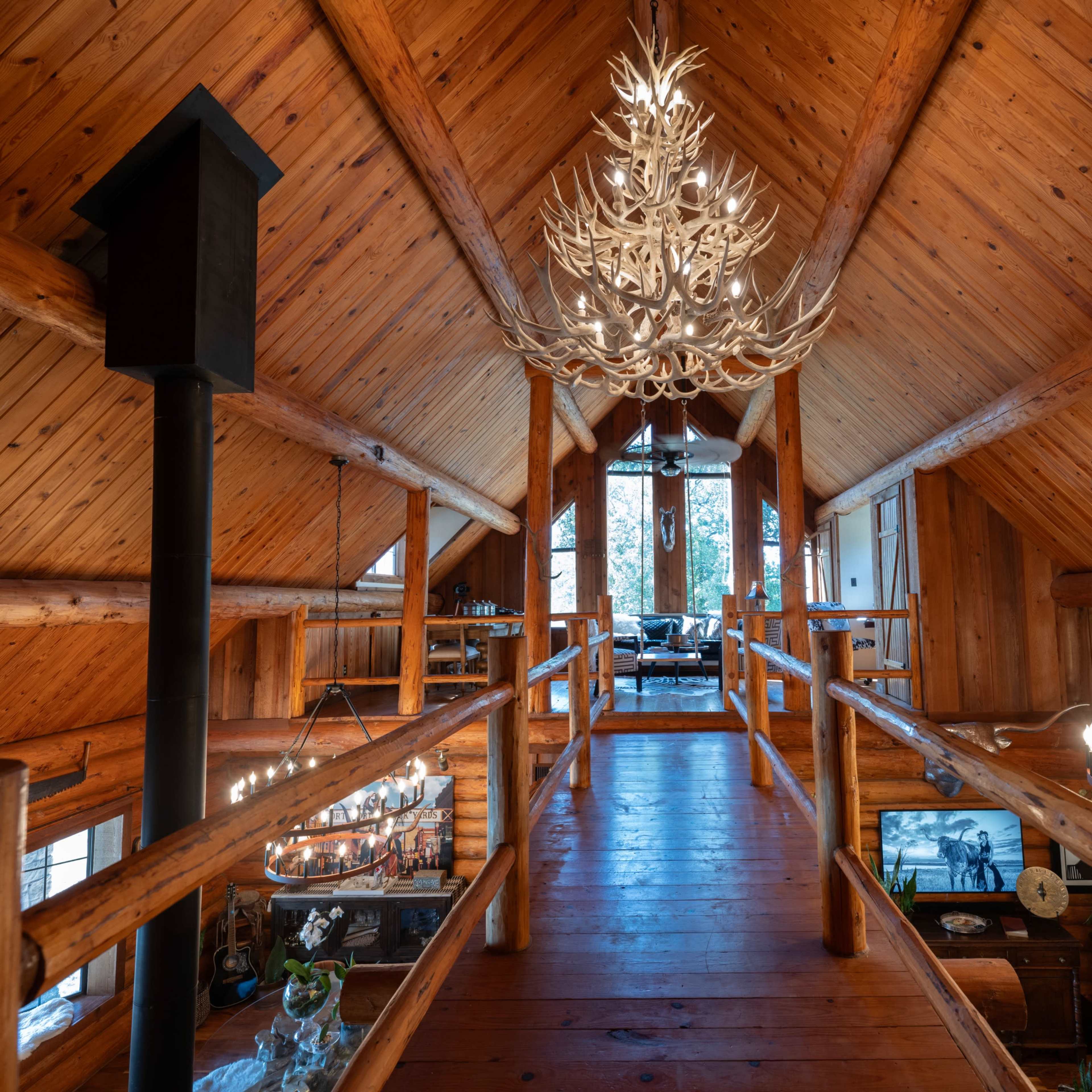A wooden loft with a high ceiling features a large antler chandelier and multiple windows overlooking a living area below.