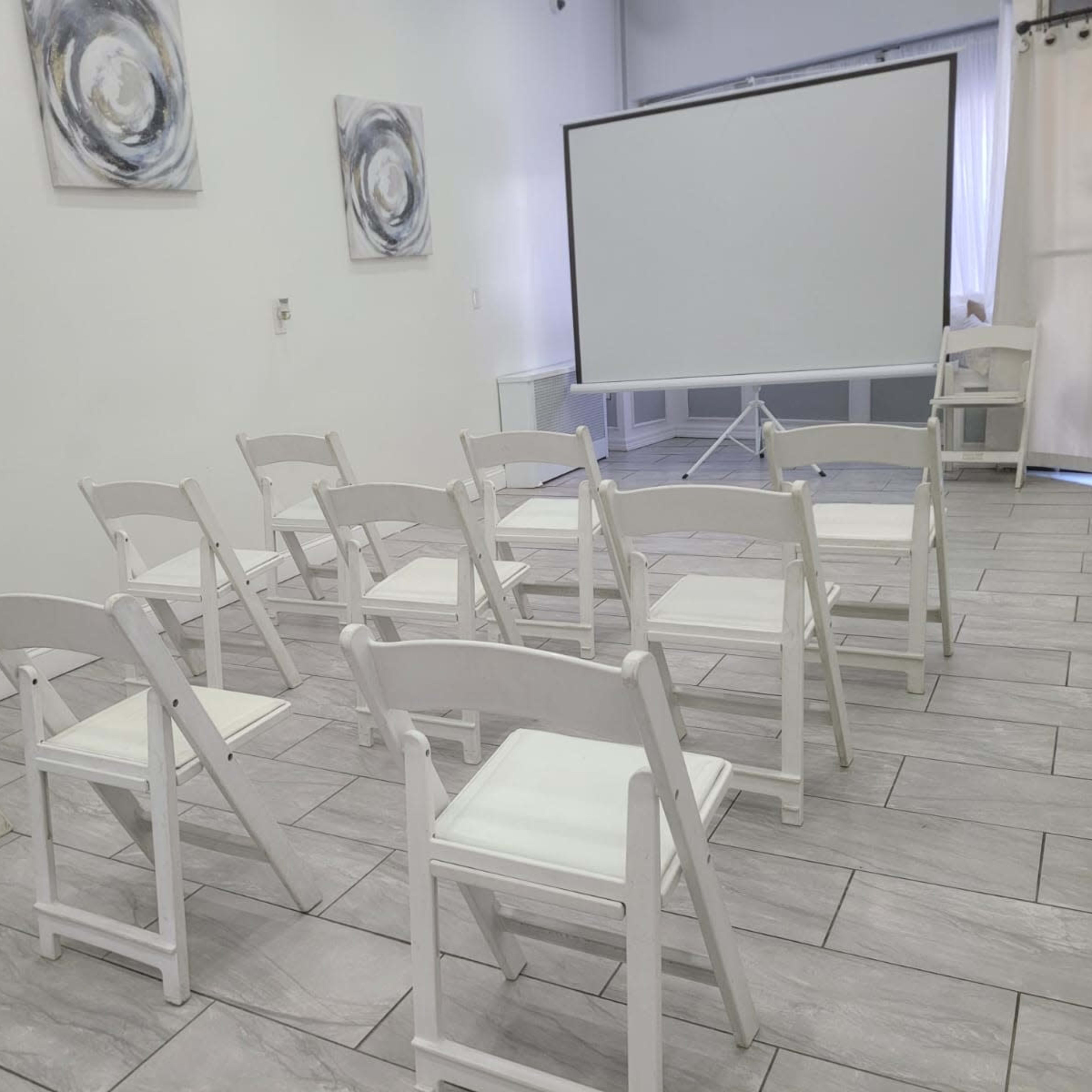 A row of white folding chairs is arranged facing a blank projection screen in a well-lit room with abstract wall art.