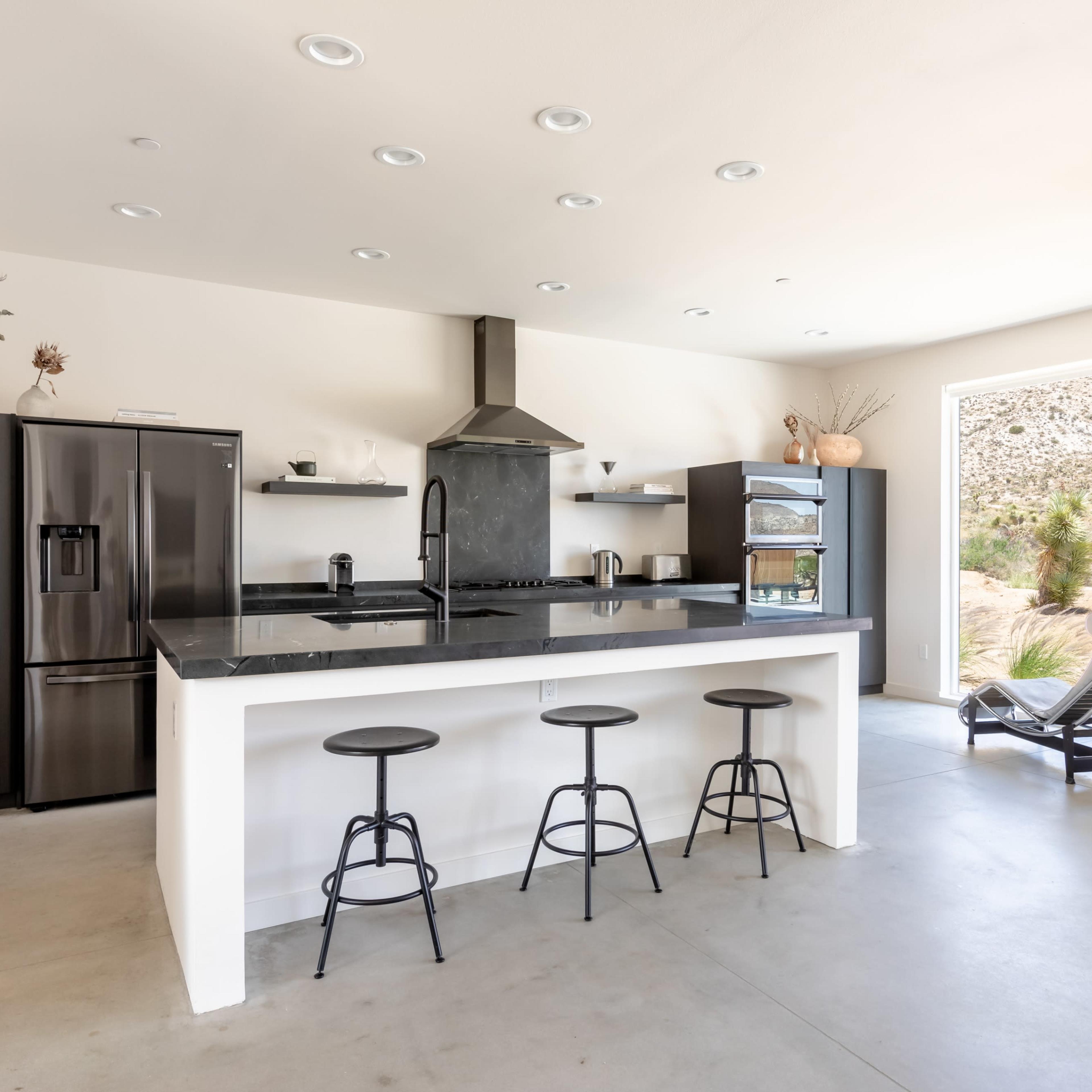 A modern kitchen features a black countertop, stainless steel appliances, and three black bar stools at a white island, with large windows overlooking a desert landscape.