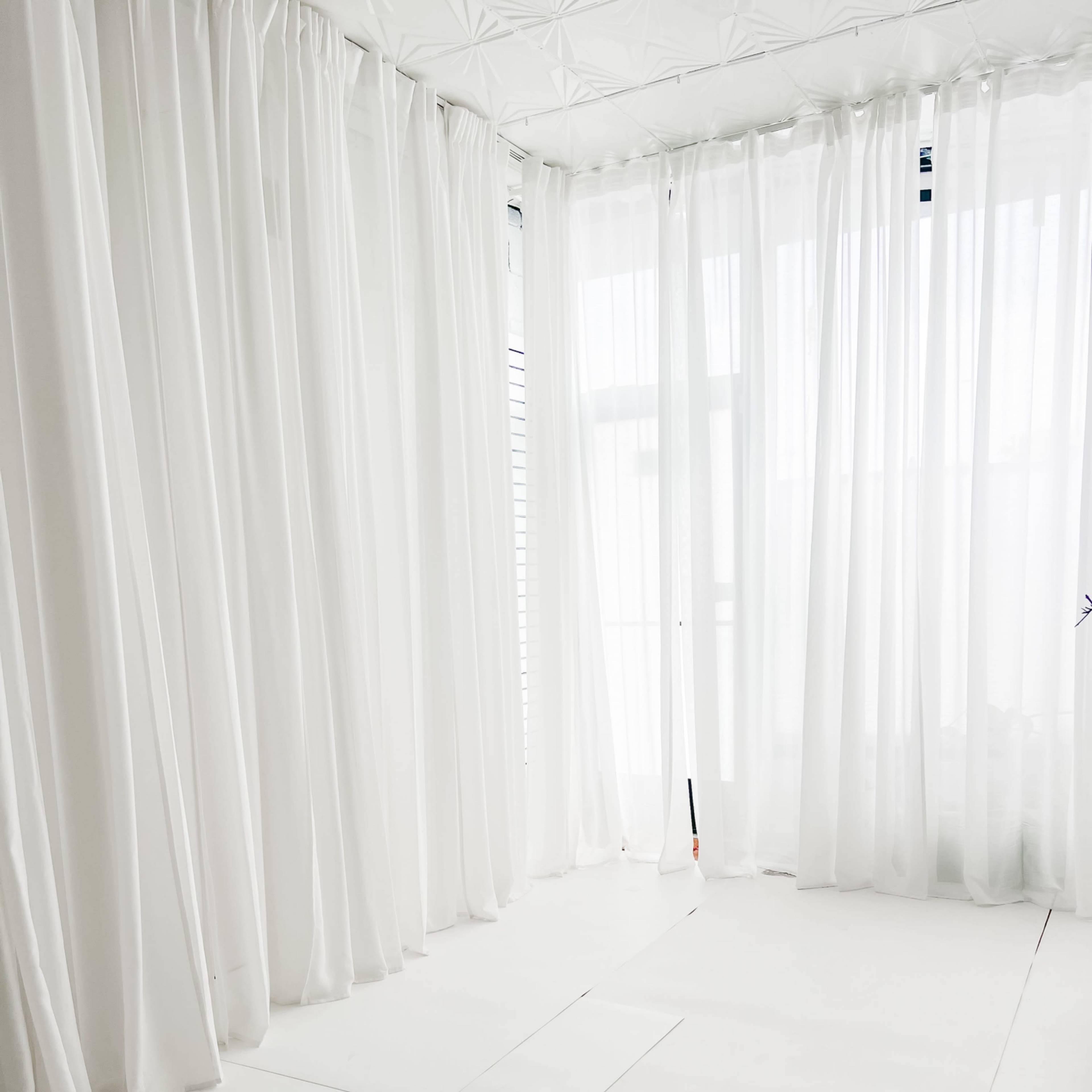 A bright, empty room features white walls and floor, with sheer white curtains allowing natural light to enter.