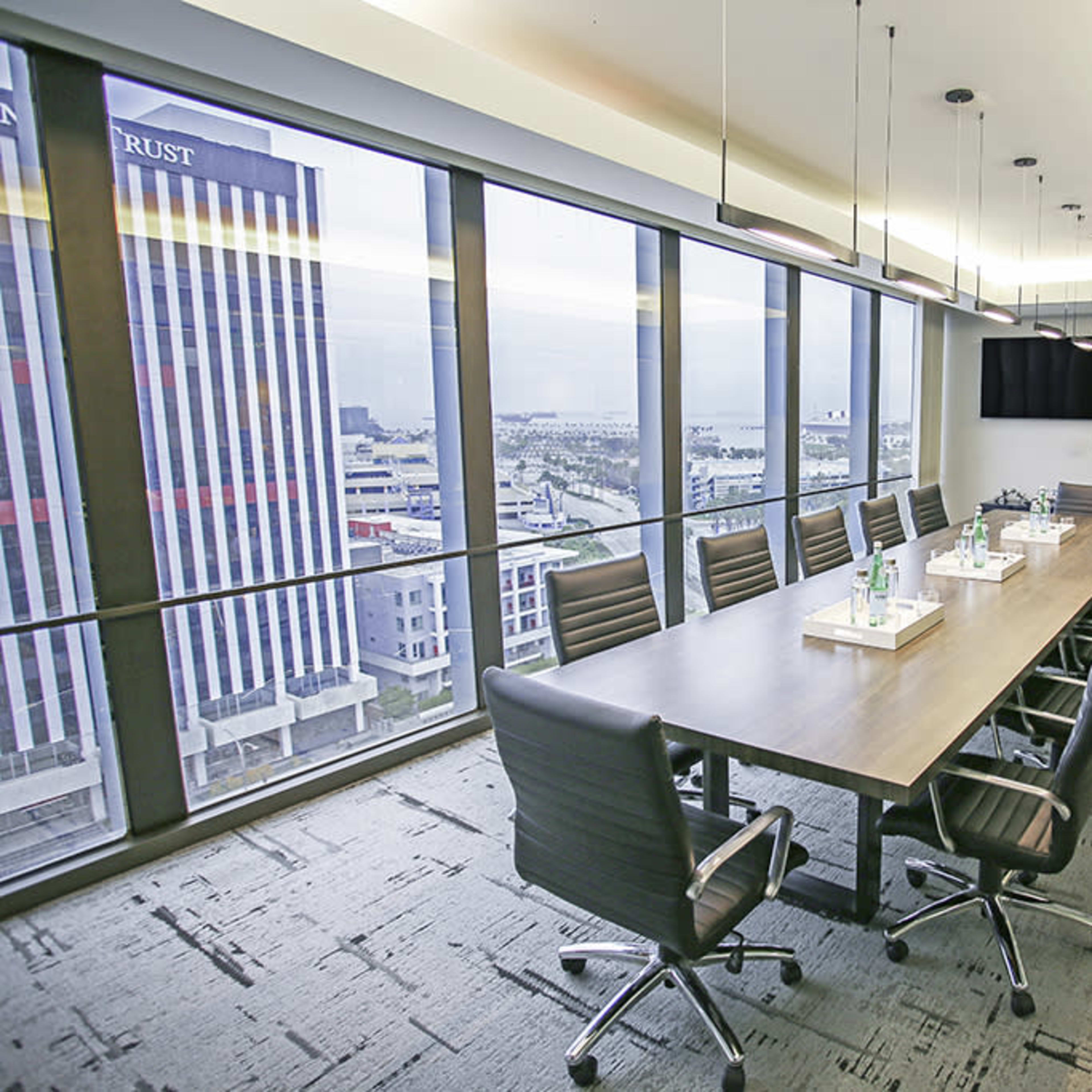 A spacious conference room features a long wooden table surrounded by ergonomic chairs, with large windows offering a view of the city skyline.