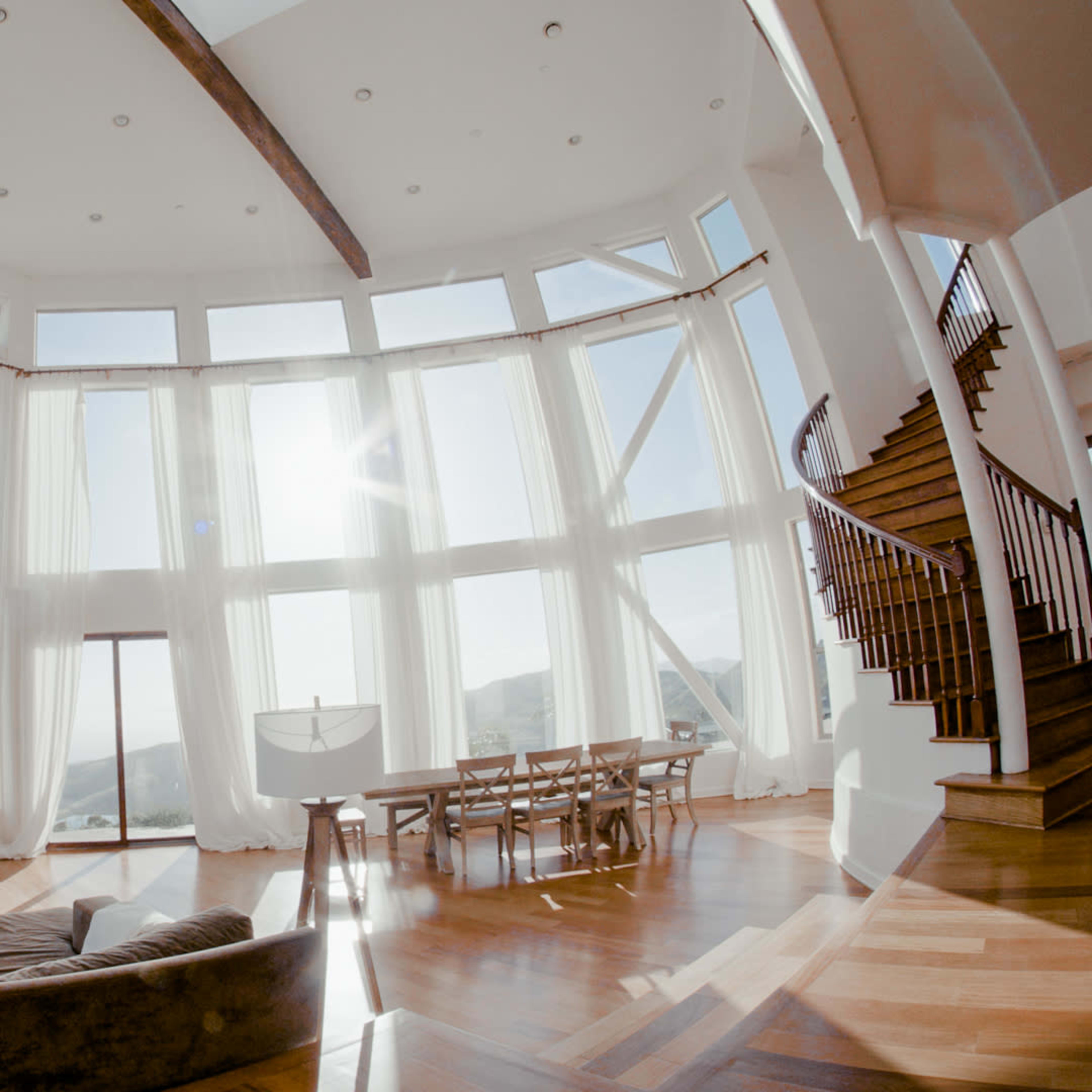 A spacious living room features large windows with sheer curtains, a staircase, a dining area, and a television.