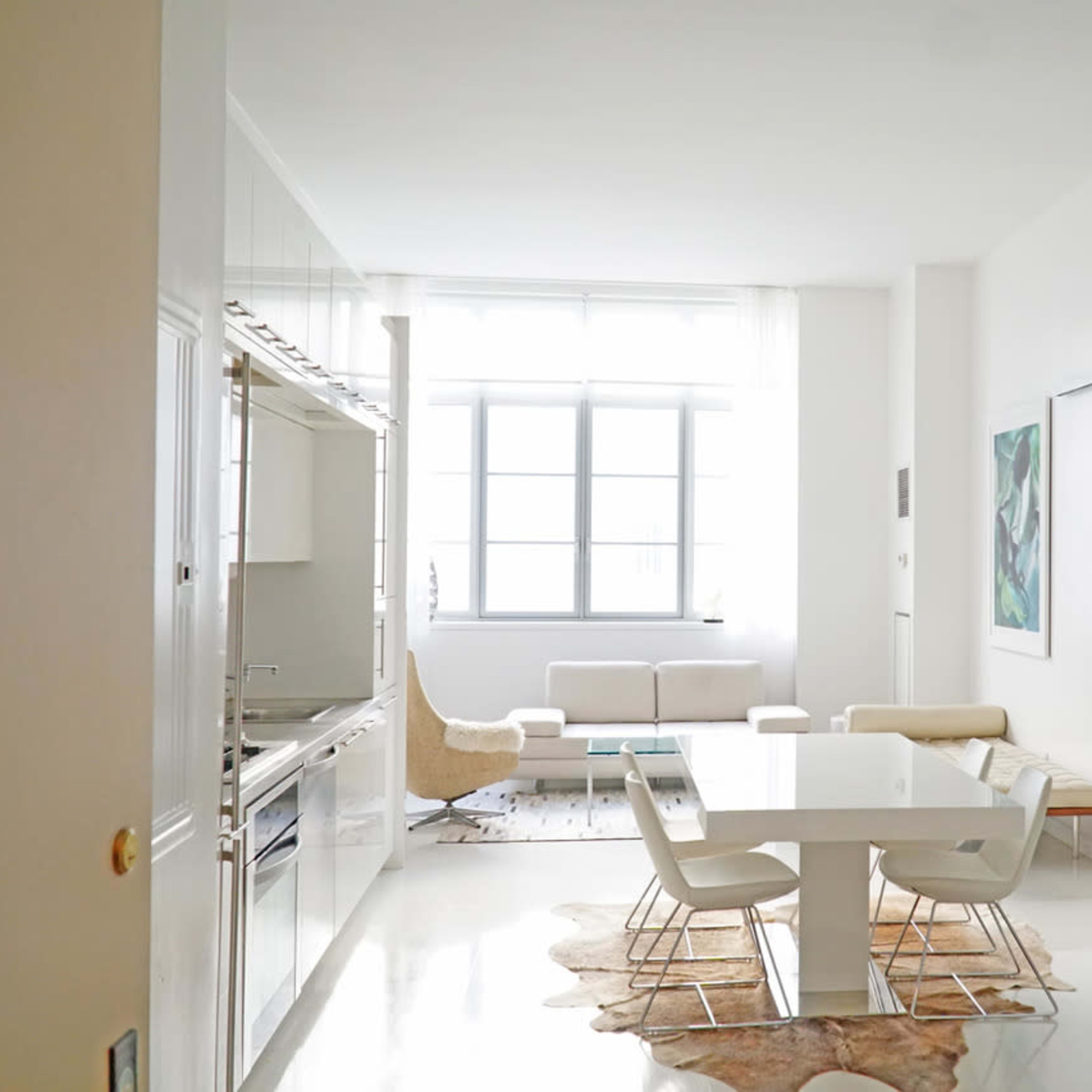 A modern kitchen and dining area with a glass wall, a white dining table, and a light-filled living space featuring a sofa and decorative artwork.