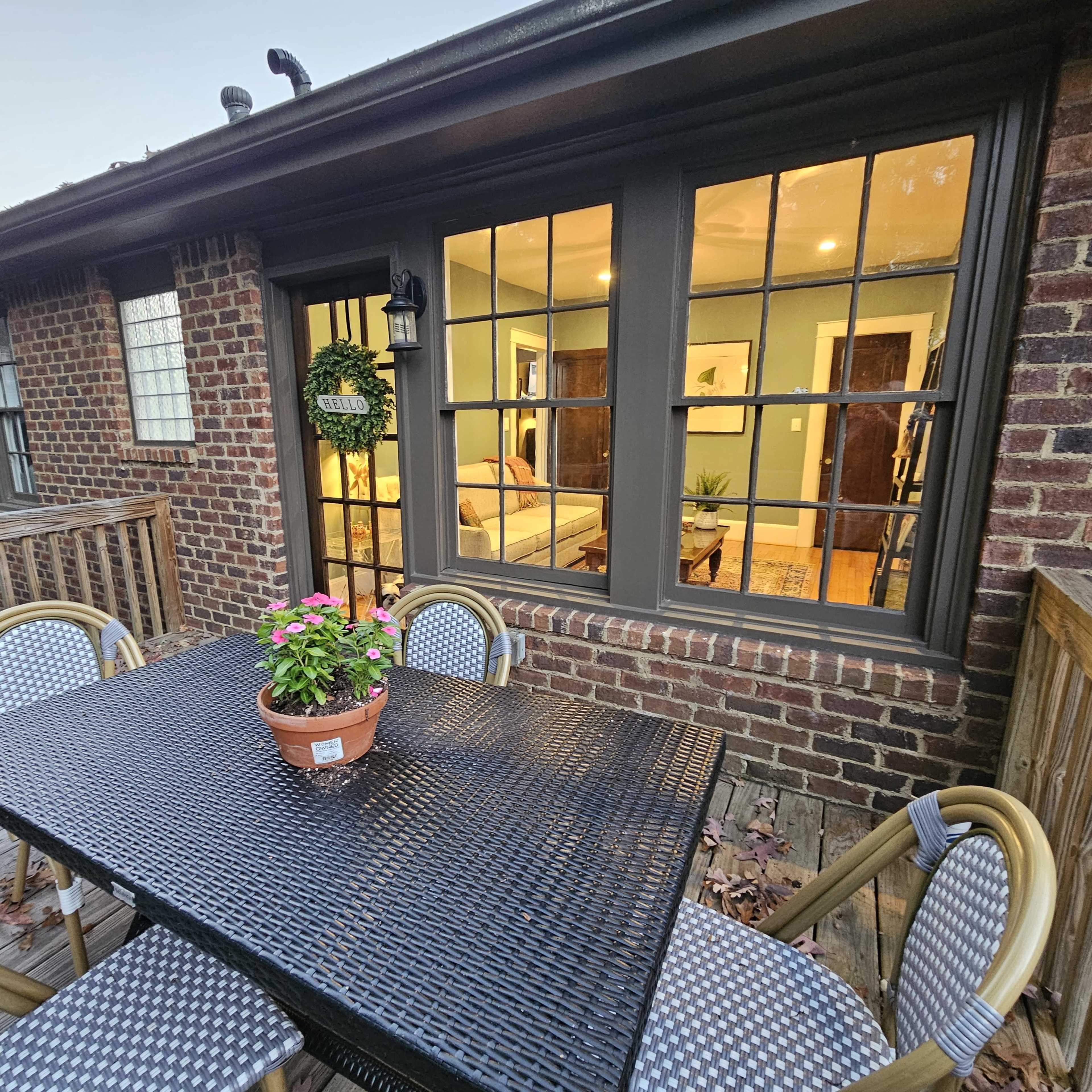 A patio table with a potted plant sits outside a brick home, showcasing a well-lit room visible through a window.