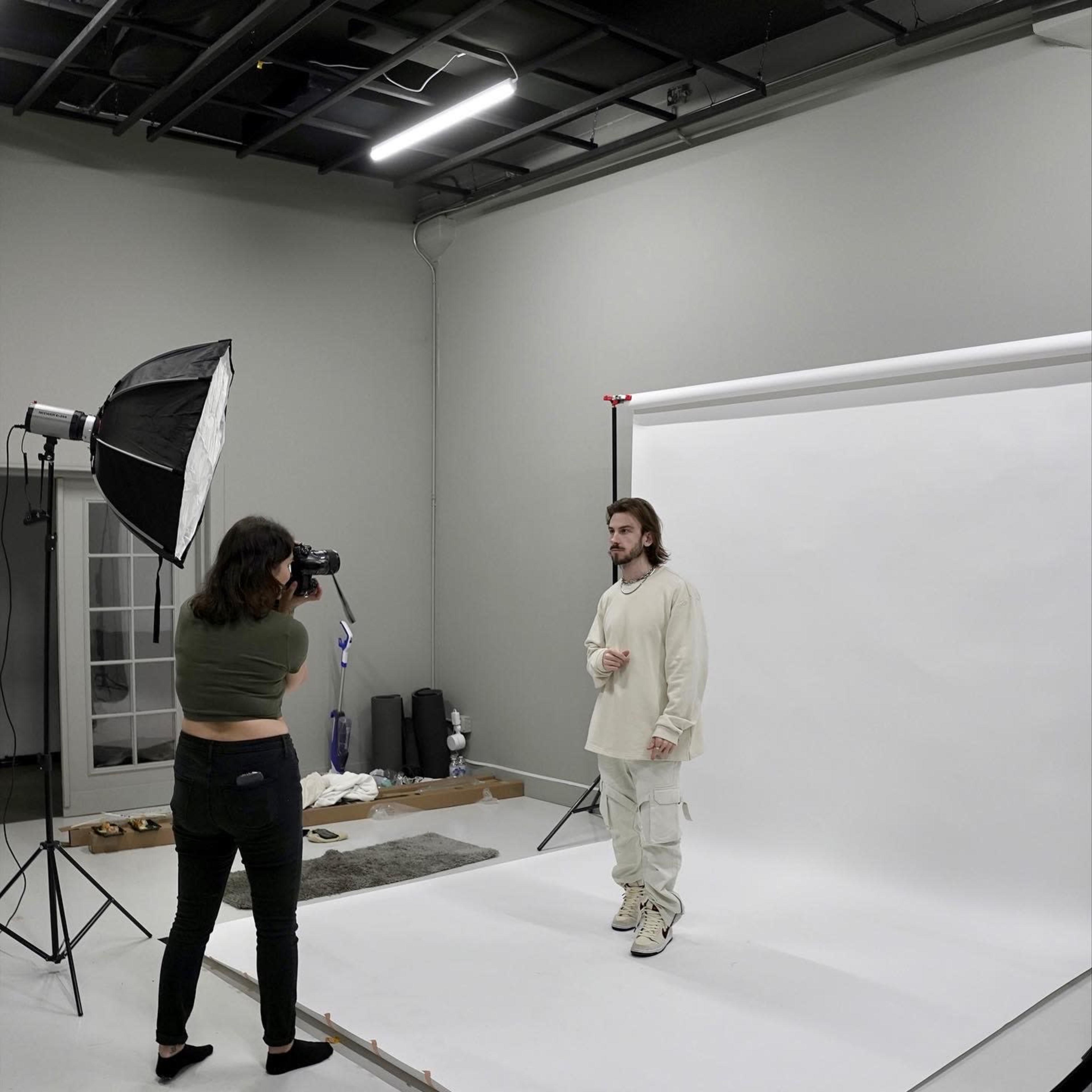 A photographer shoots a subject wearing light clothing against a plain white backdrop in a studio setting.