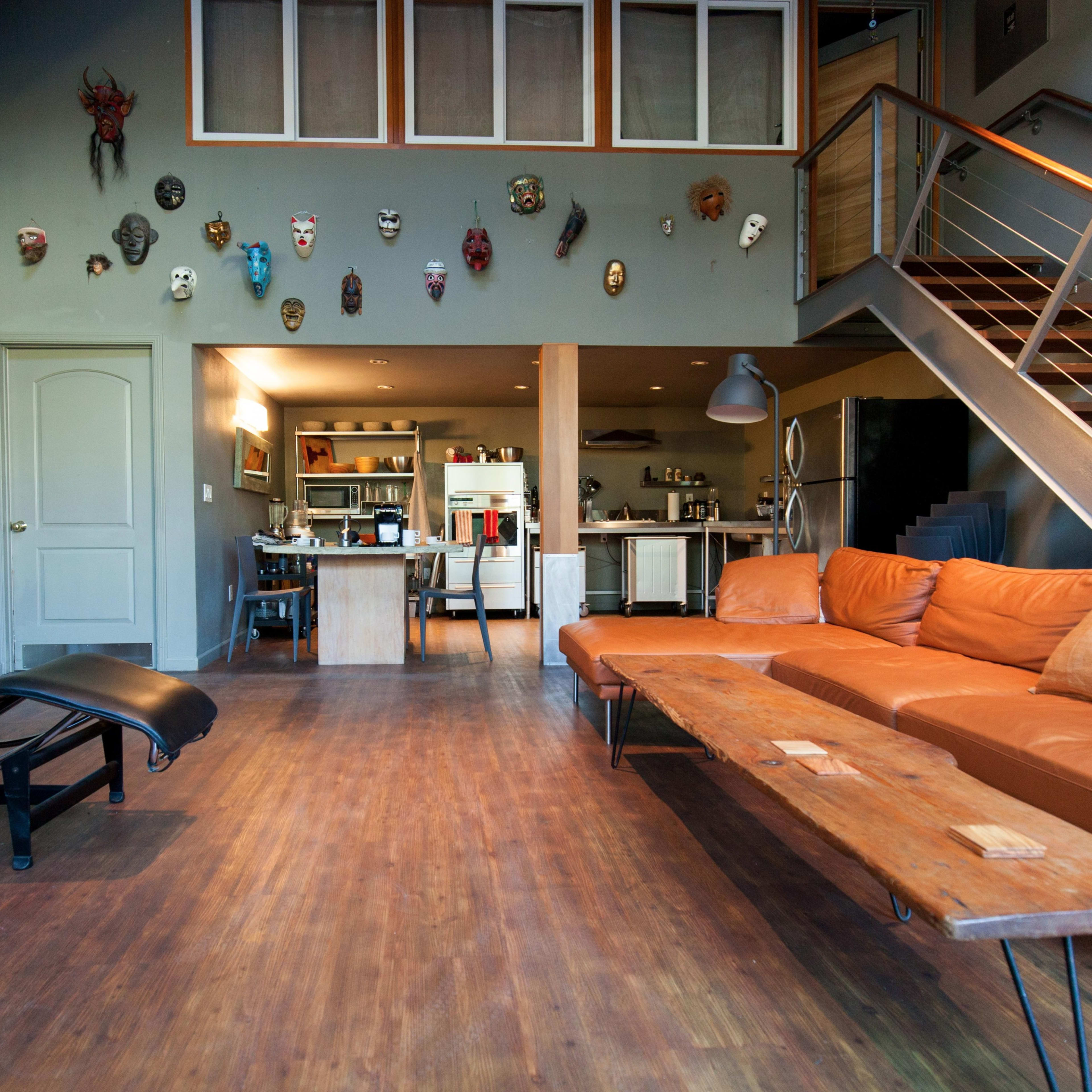 A spacious living area with a leather sofa, a chaise lounge, and a wooden coffee table, featuring eclectic wall decor and an open kitchen in the background.