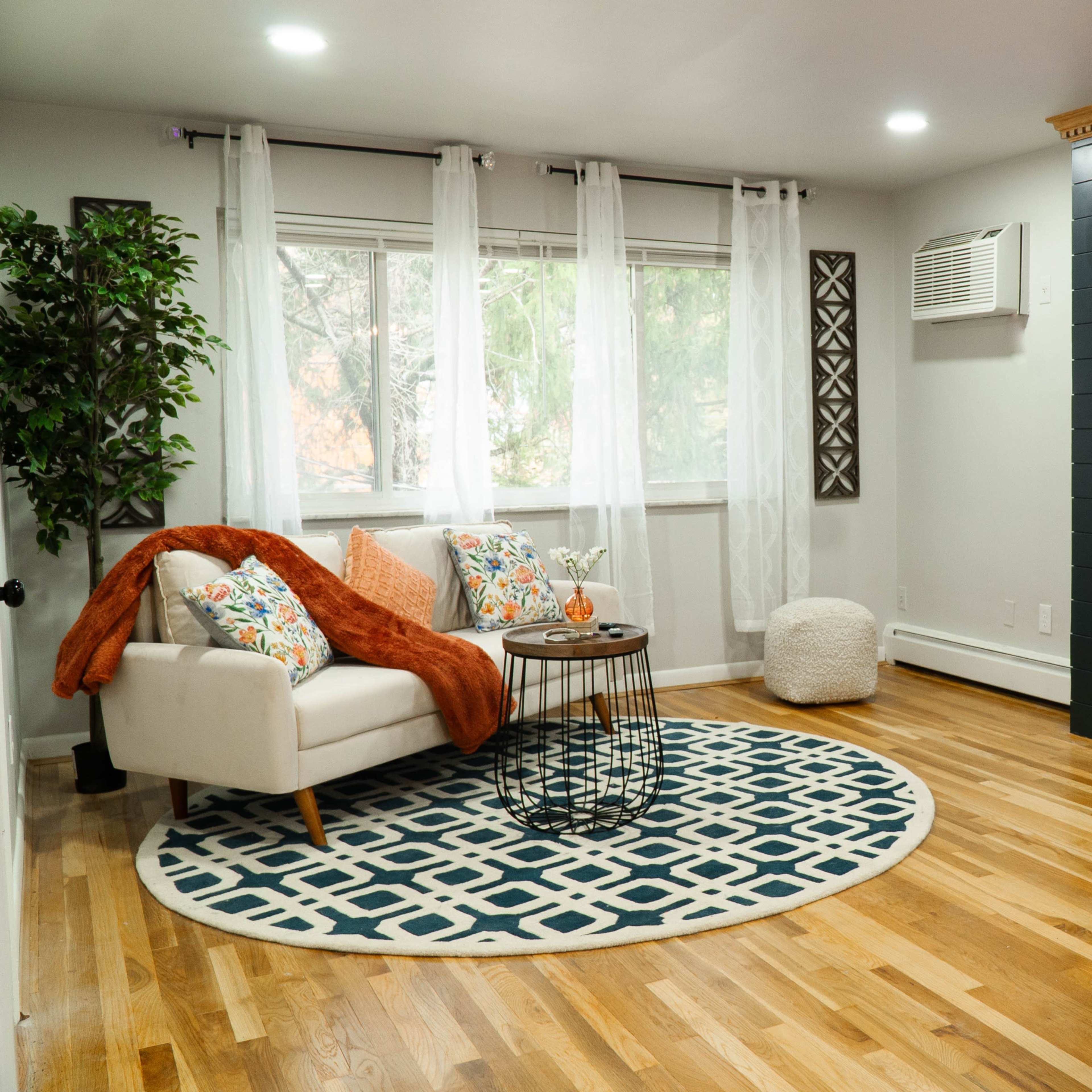 A modern living room features a white couch with colorful pillows, a round rug, a wall-mounted television, and an electric fireplace.