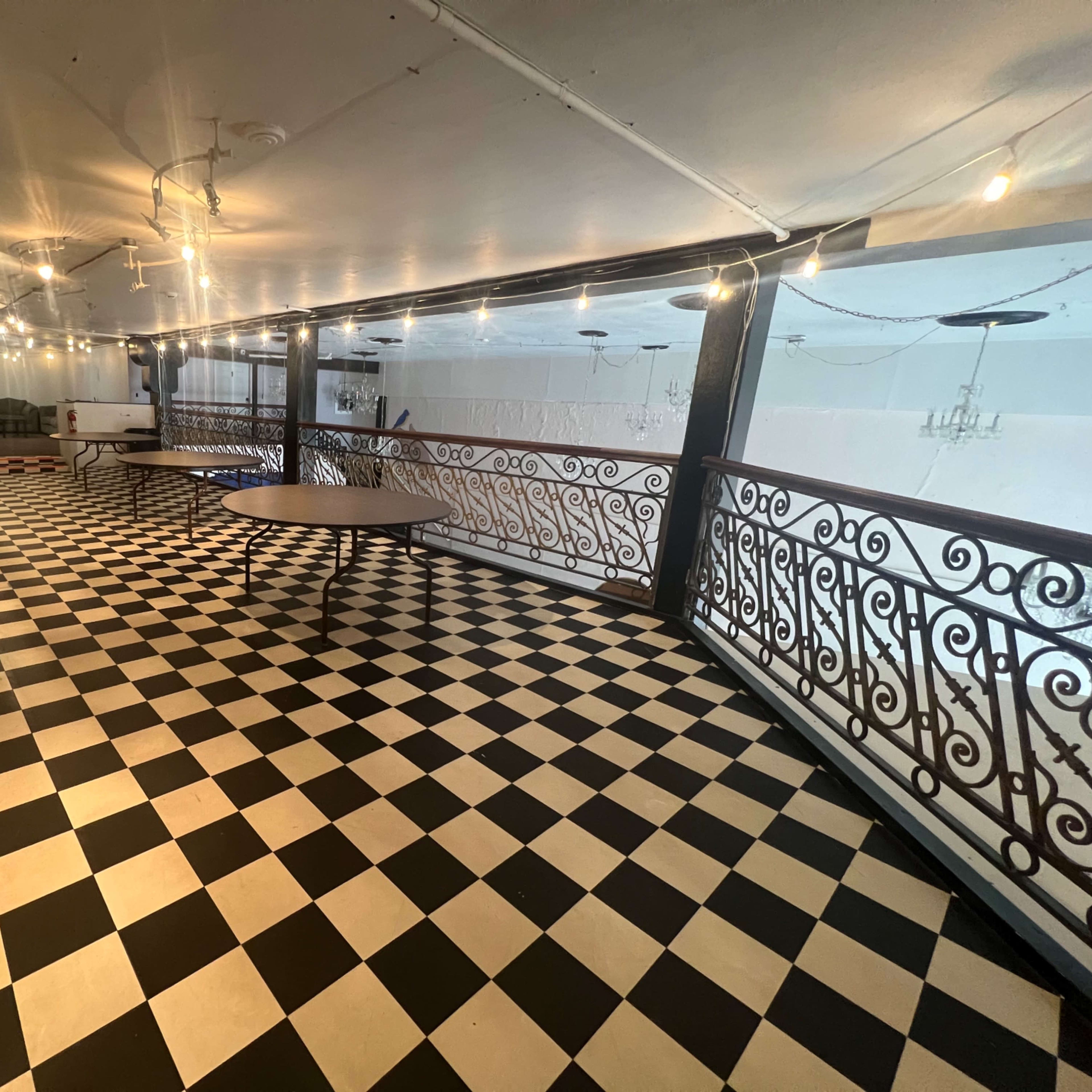 A spacious balcony area with a black and white checkered floor, decorative iron railings, and overhead string lights.