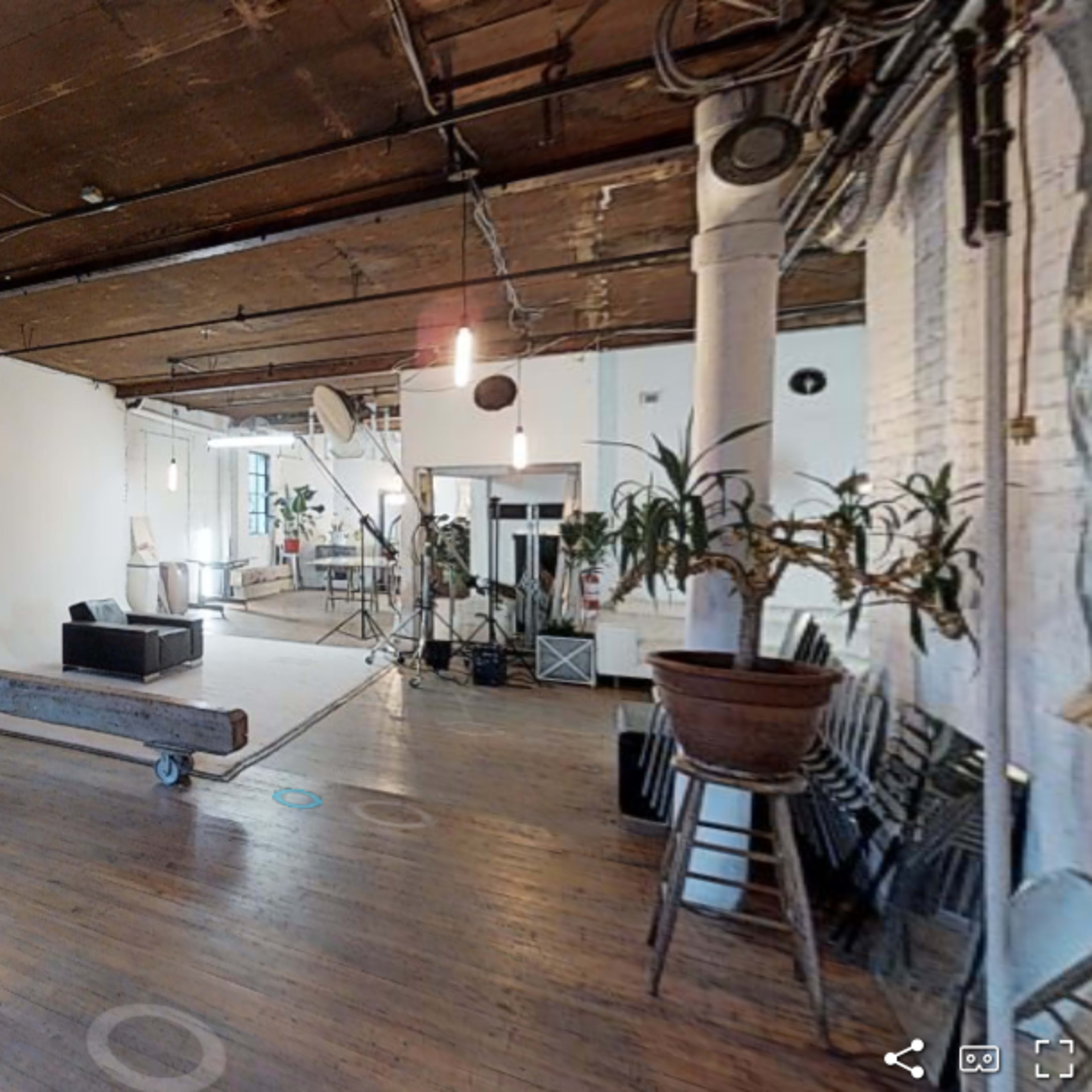 An industrial-style loft space with wooden floors, a large window, potted plants, a sofa, and a minimalist decor featuring a staircase in the background.