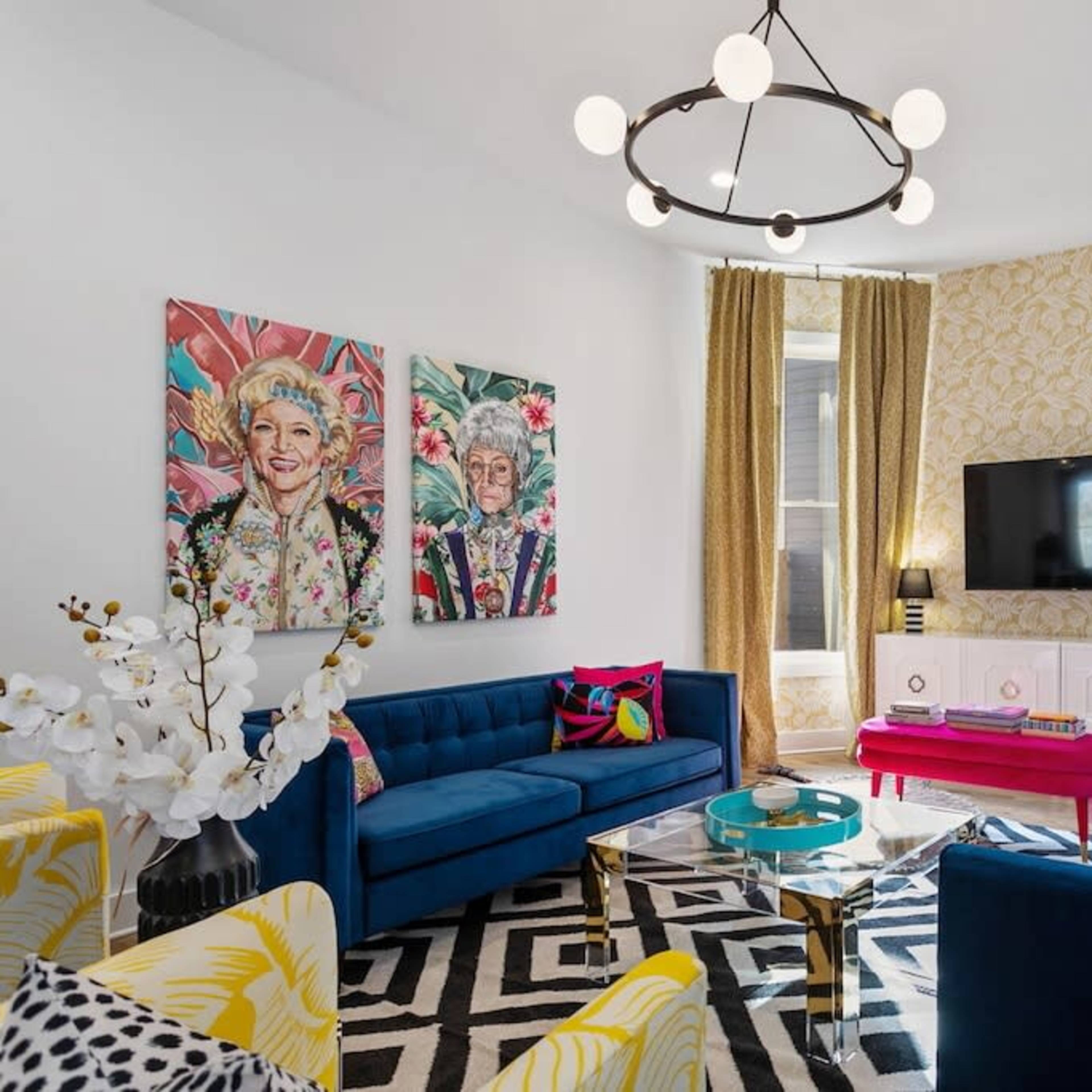 A brightly decorated living room featuring blue sofas, colorful cushions, patterned curtains, and wall art, with a television mounted above a white cabinet.