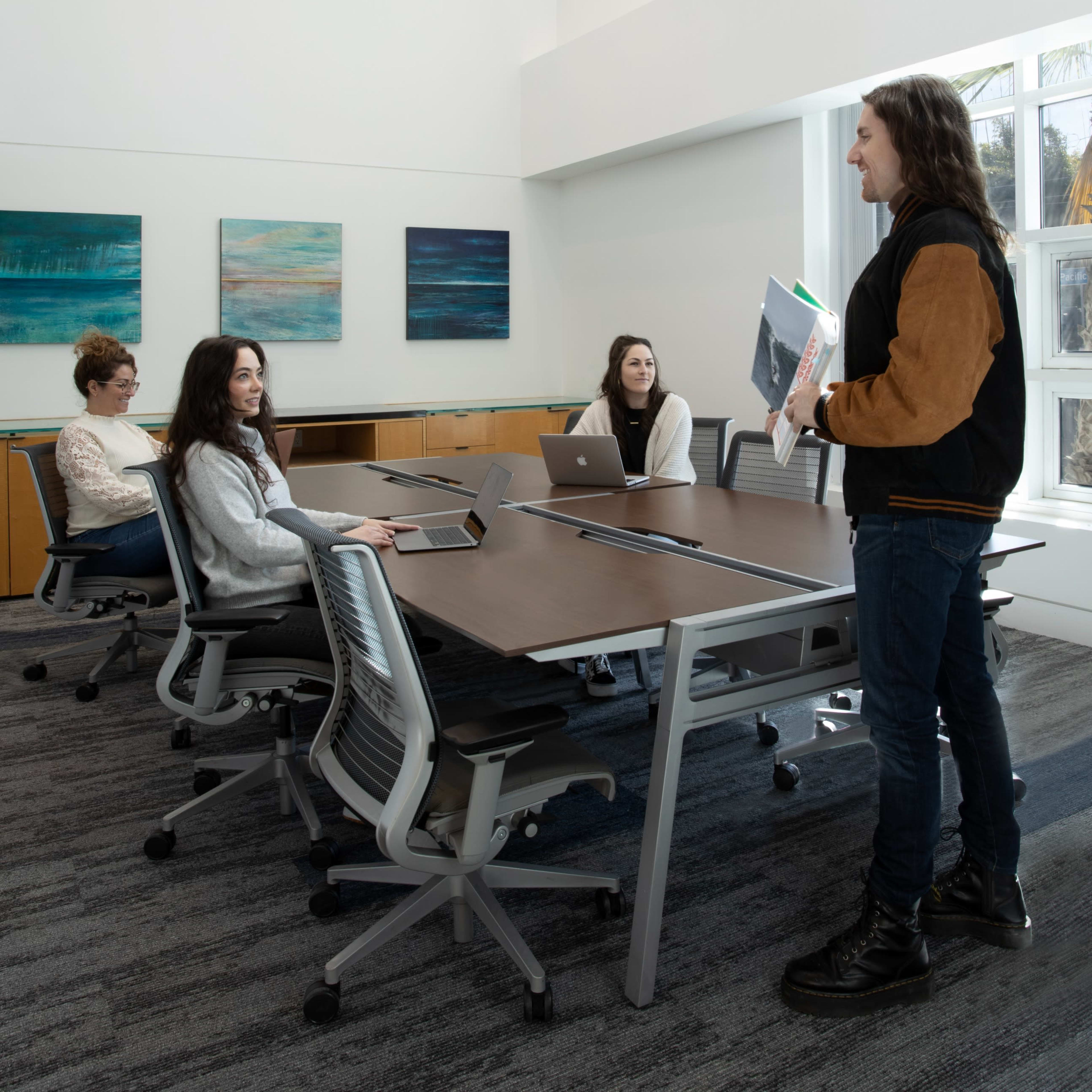 A group of four people are engaged in a discussion around a conference table in a modern meeting room with large windows.