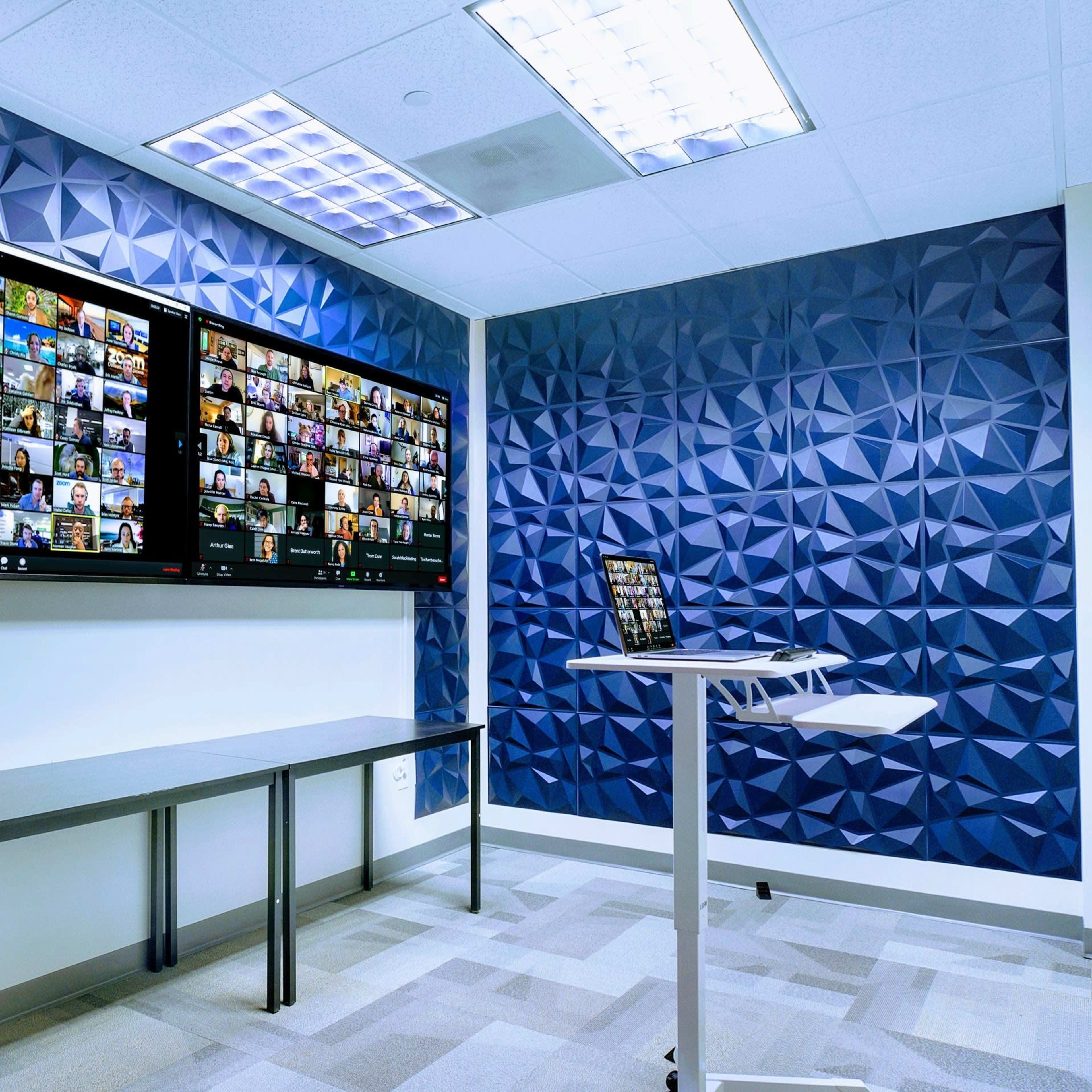 A modern conference room with two large screens displaying a Zoom meeting and a small table with a laptop.