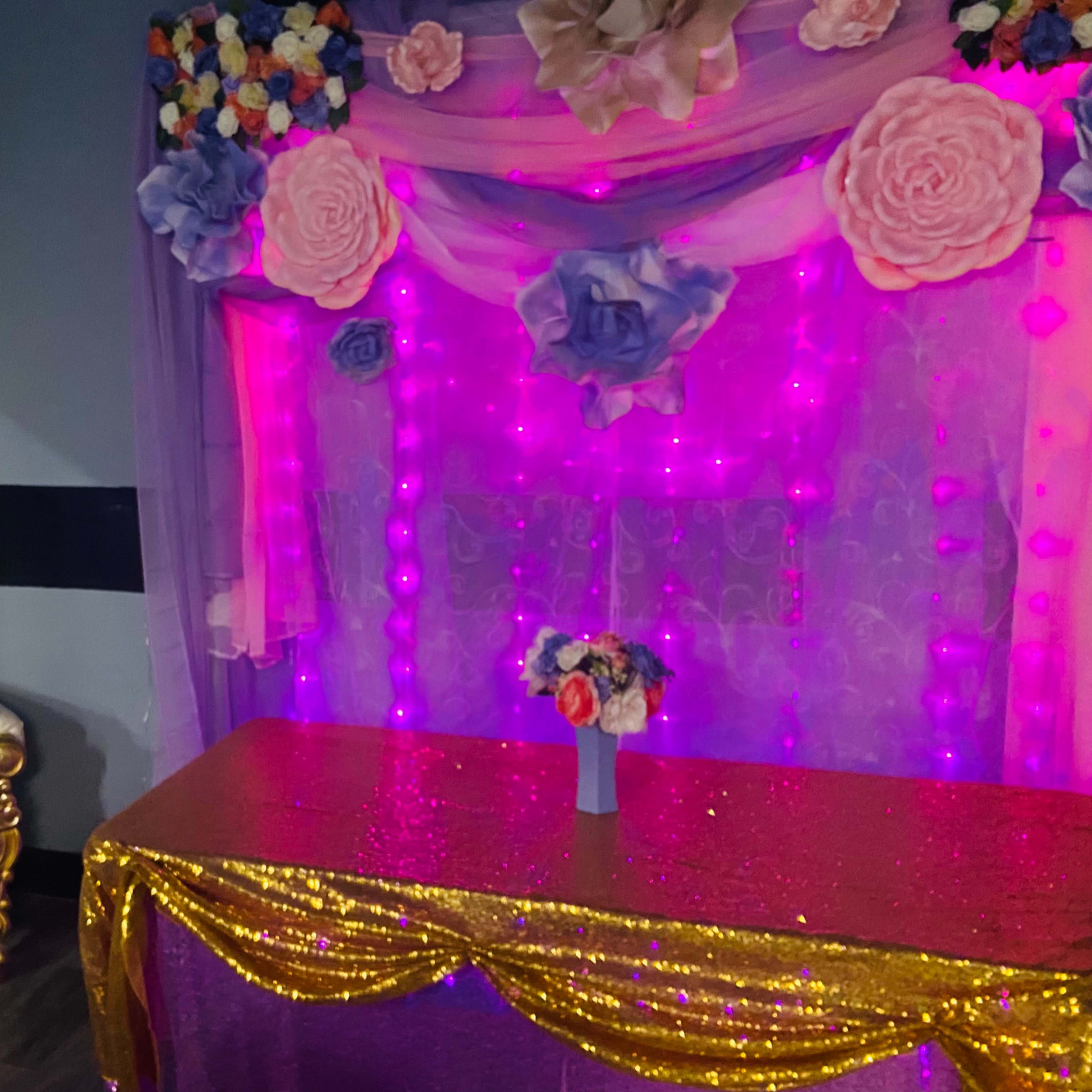 A decorative setup featuring a gold-accented chair, a table draped in shiny purple fabric, and a backdrop of lights and floral decorations.