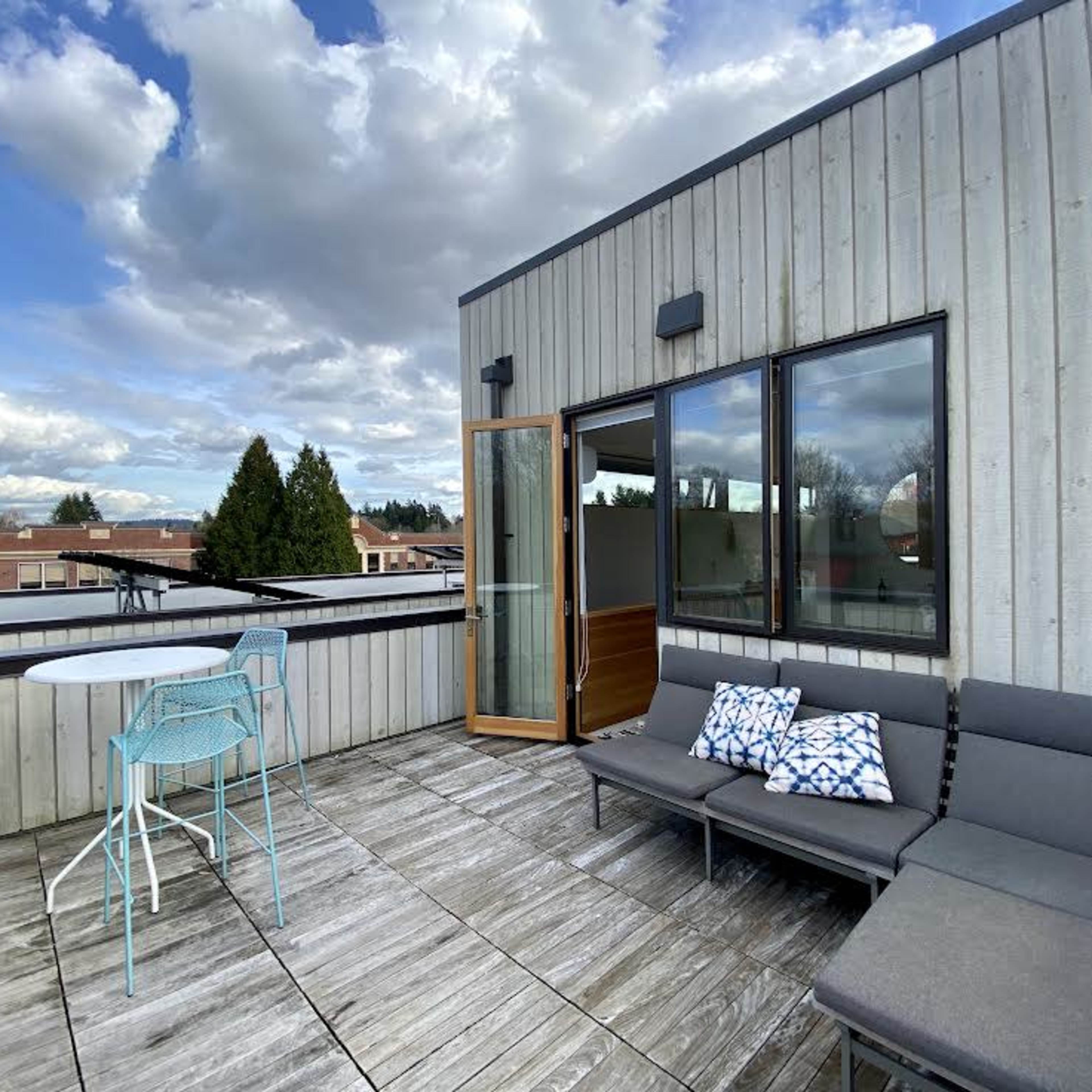 A rooftop deck with a small table and chairs, a lounge area, and large windows leading to an interior space.