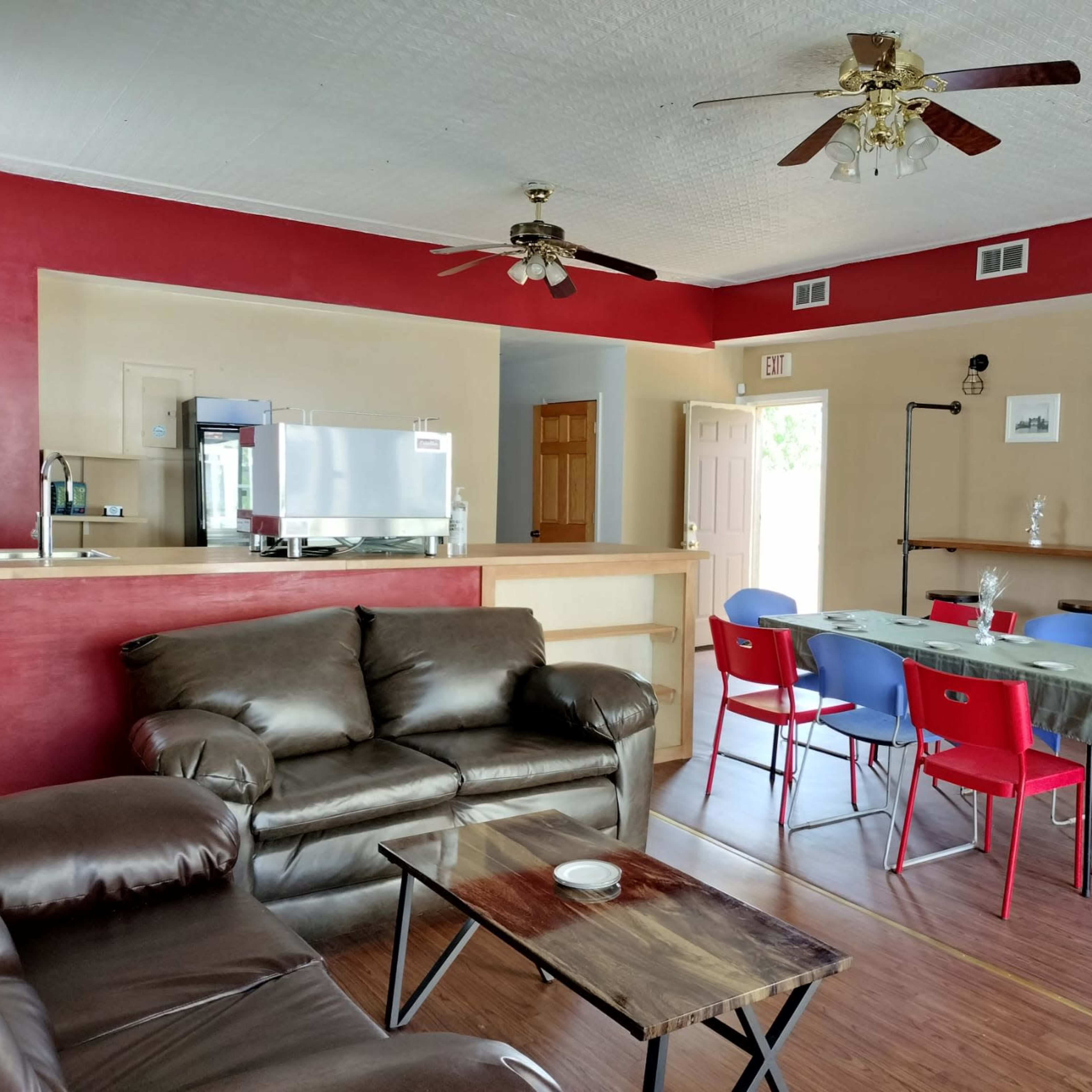 A living room with a brown leather couch, a wooden table, red and blue chairs, and a kitchen area in the background.