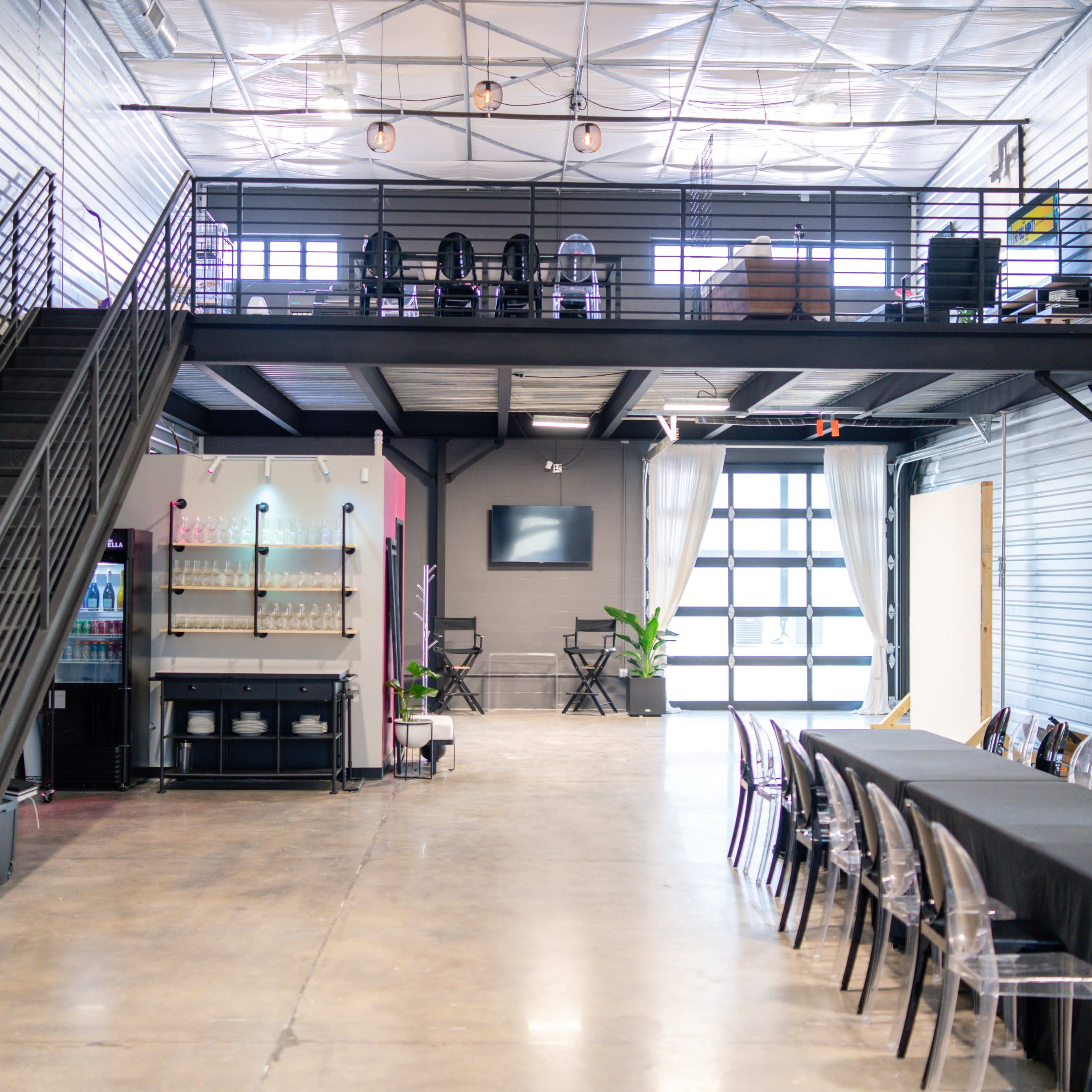 A spacious indoor venue with a black staircase leading to a mezzanine, a long table set up on the ground floor, and a bar area in the background.