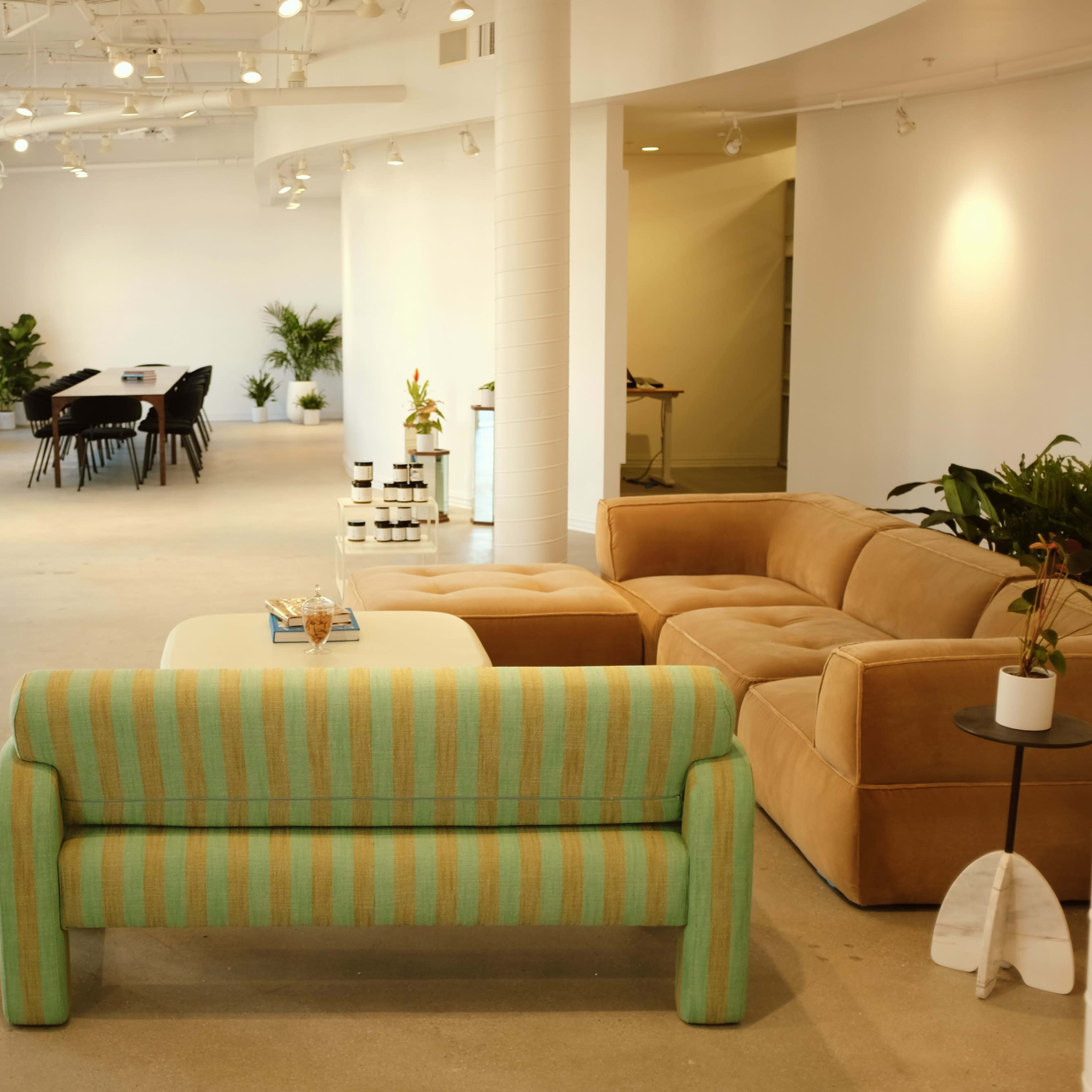 A spacious, modern office interior with a striped couch, a coffee table, and a dining area in the background featuring a long table and chairs.
