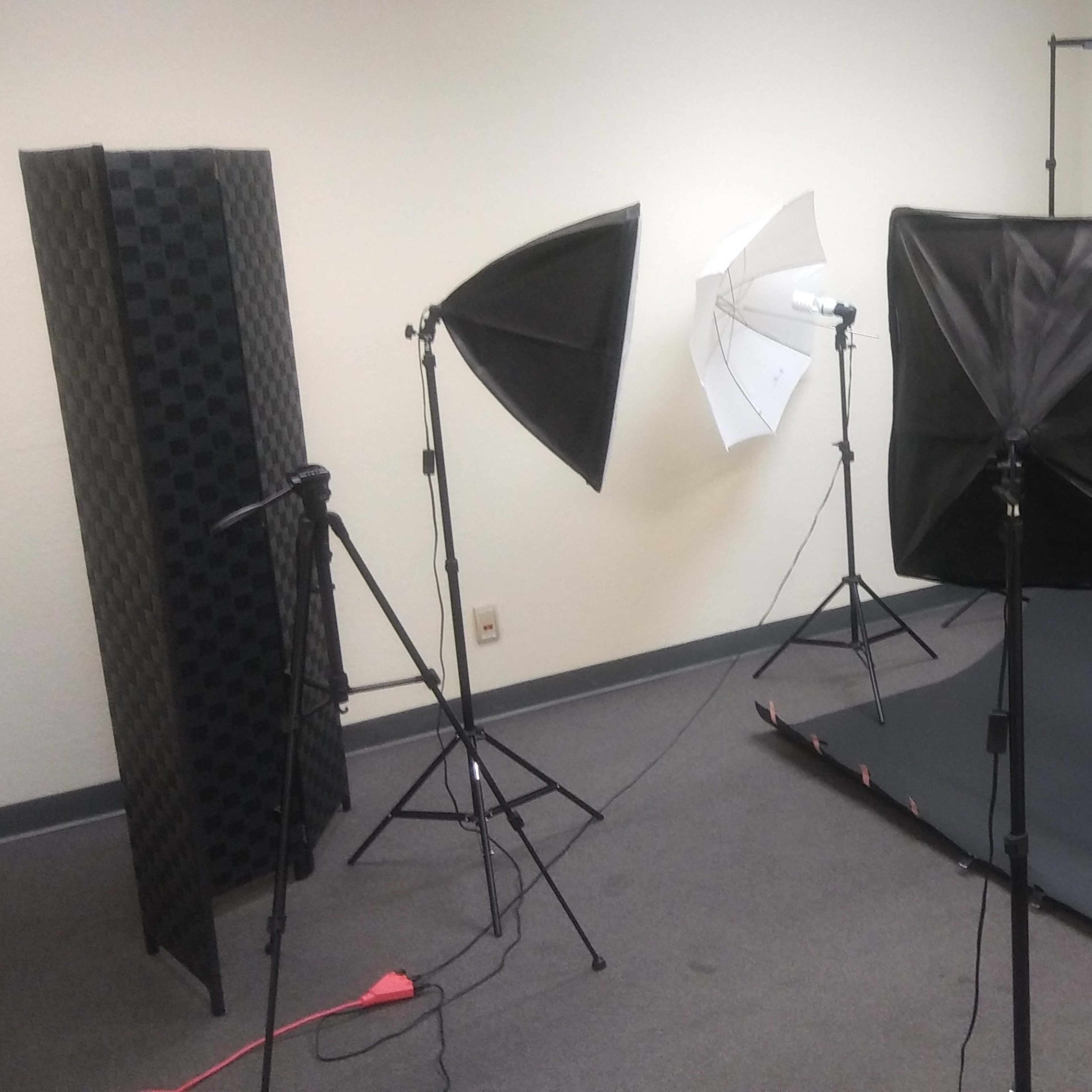 A photography studio setup with three lighting softboxes, a microphone stand, and a wooden stool in front of a dark backdrop.