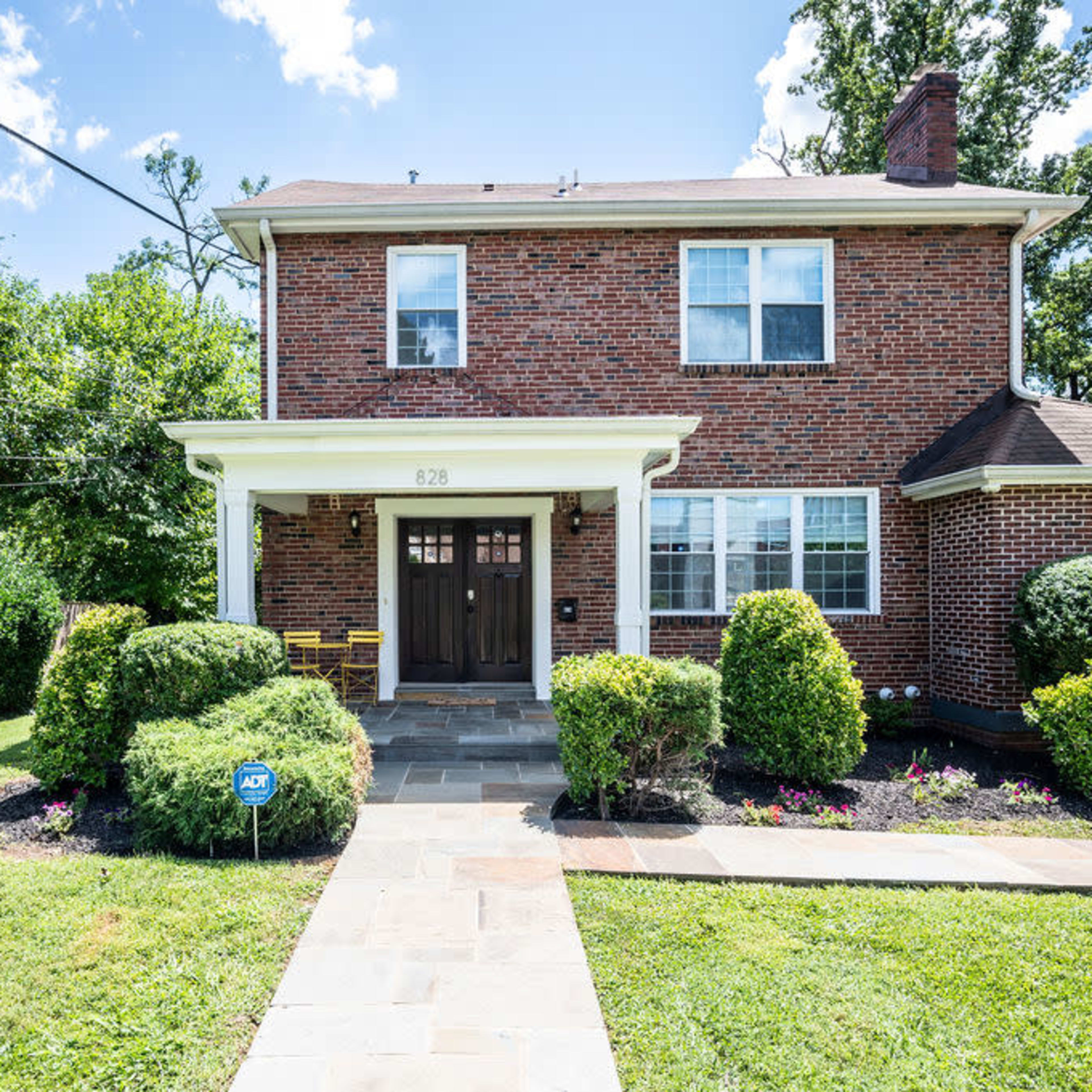 A two-story brick house with a porch, surrounded by neatly landscaped lawns and shrubs.