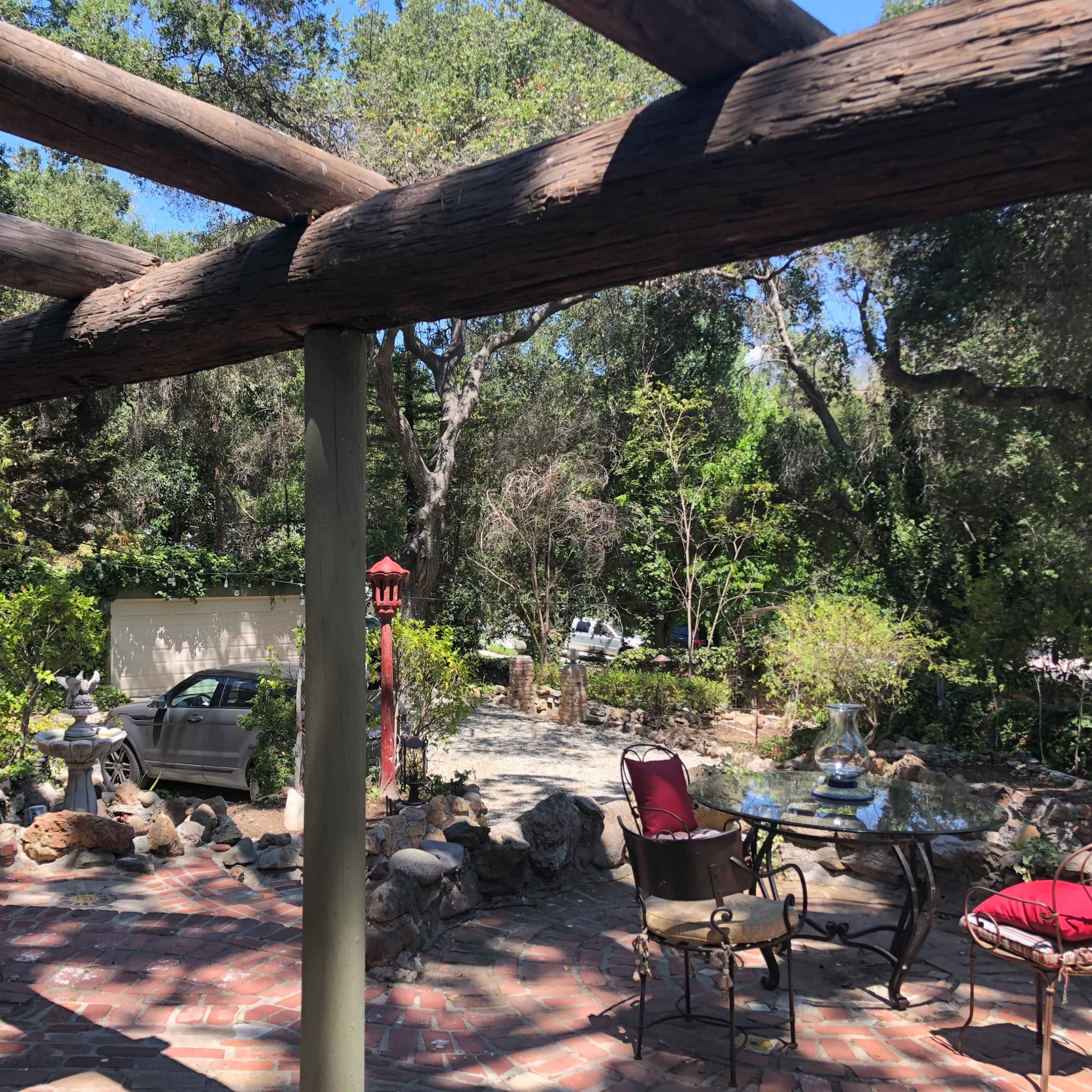 A stone patio is framed by wooden beams, overlooking a garden area with various plants and a parked car in the background.