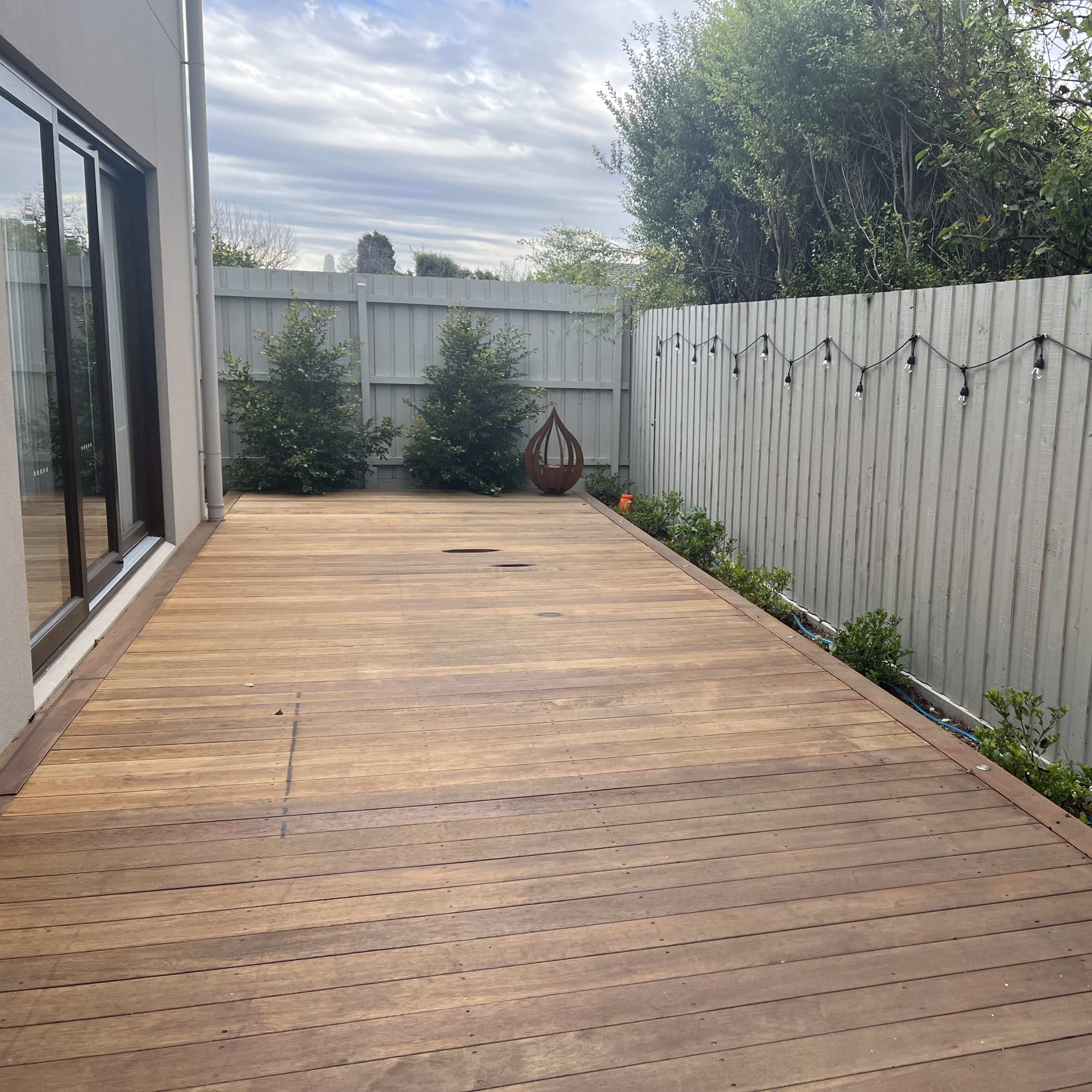 A wooden deck enclosed by a gray fence, with potted plants along one side and decorative string lights overhead.