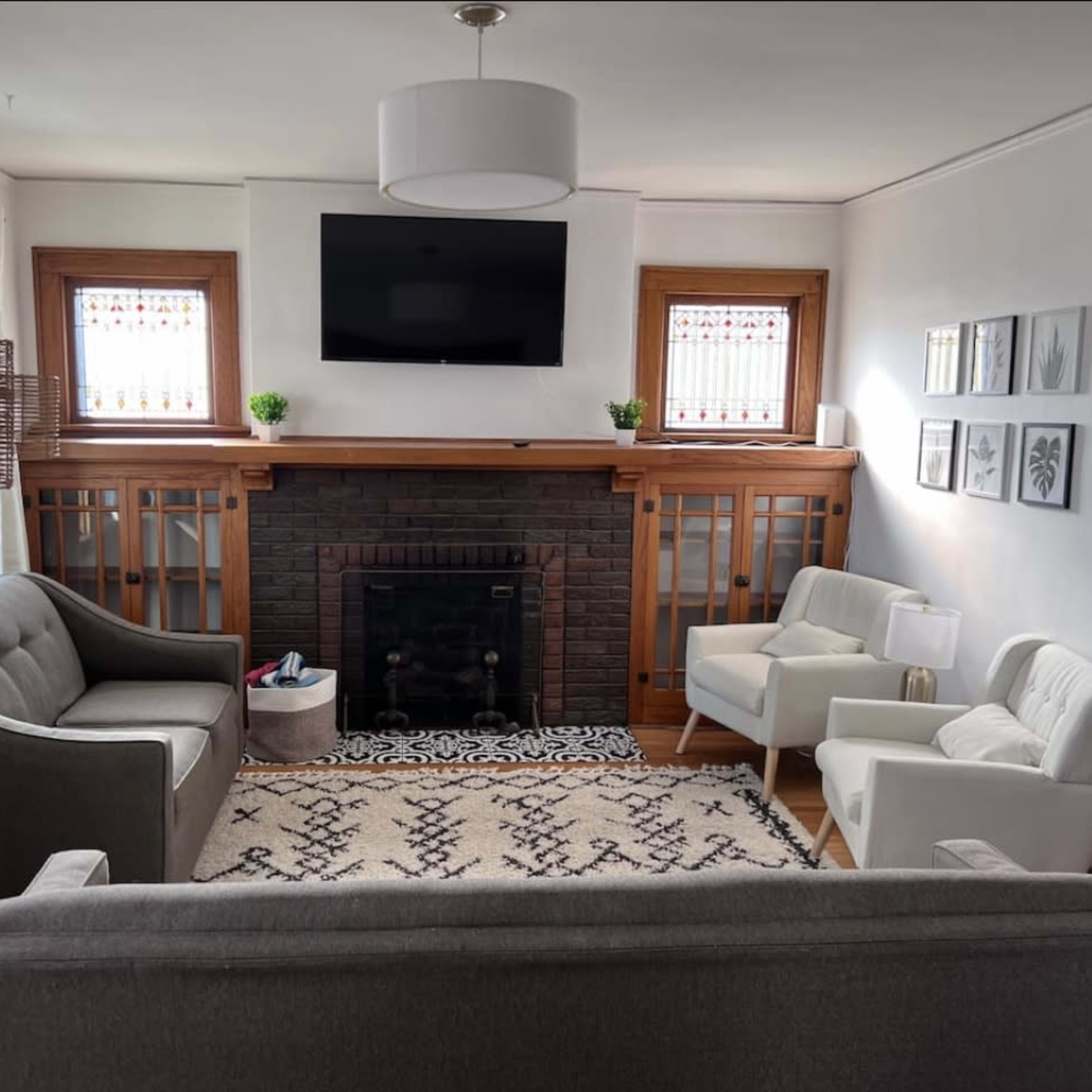 A living room features a gray sofa, two white armchairs, a black fireplace with a television above it, and decorative plants on the shelves.
