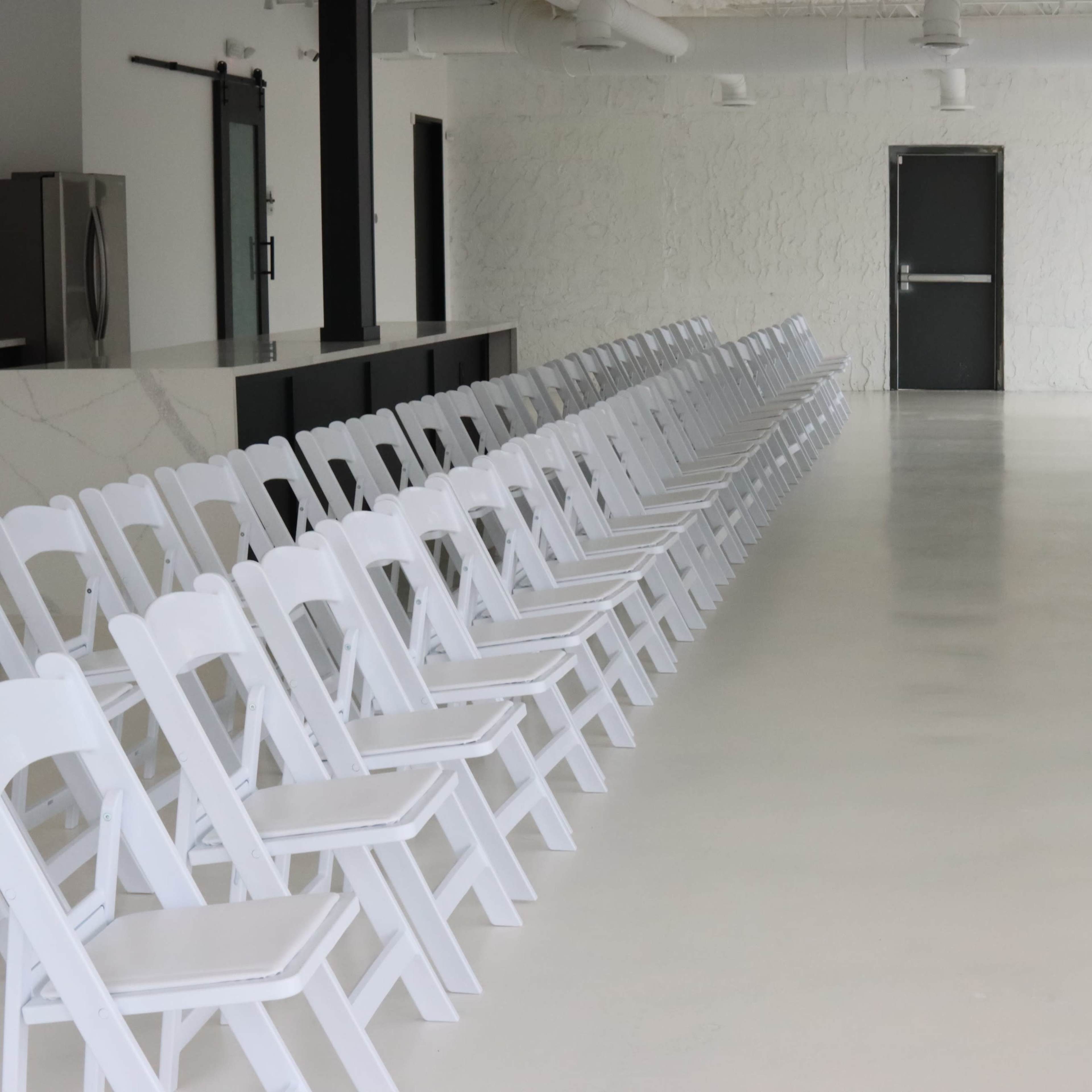A long, empty room with two rows of white folding chairs facing each other, creating a pathway in the center.