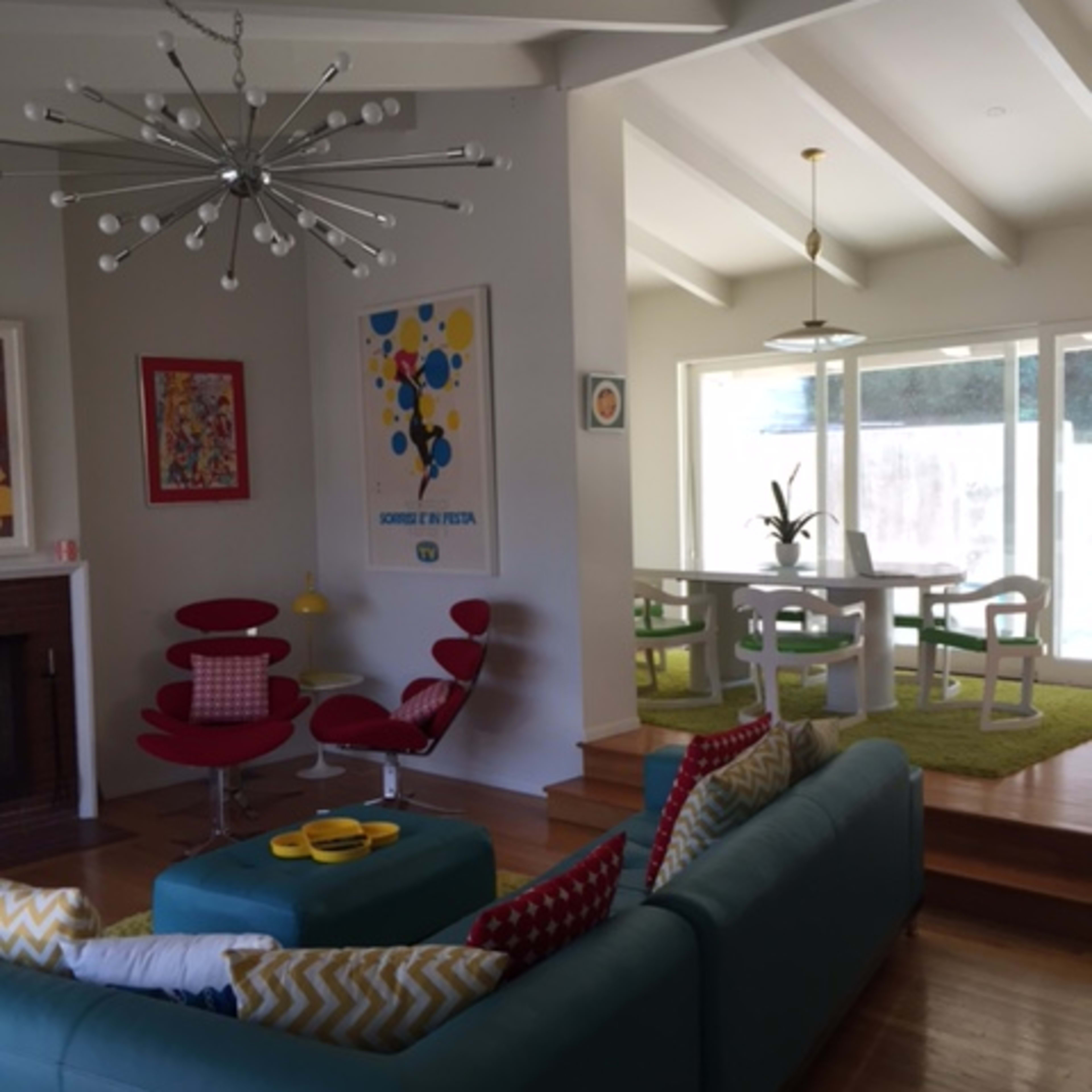 A modern living room with a turquoise sofa, red chairs, and a dining area featuring a table and chairs, accented by large windows and colorful artwork.