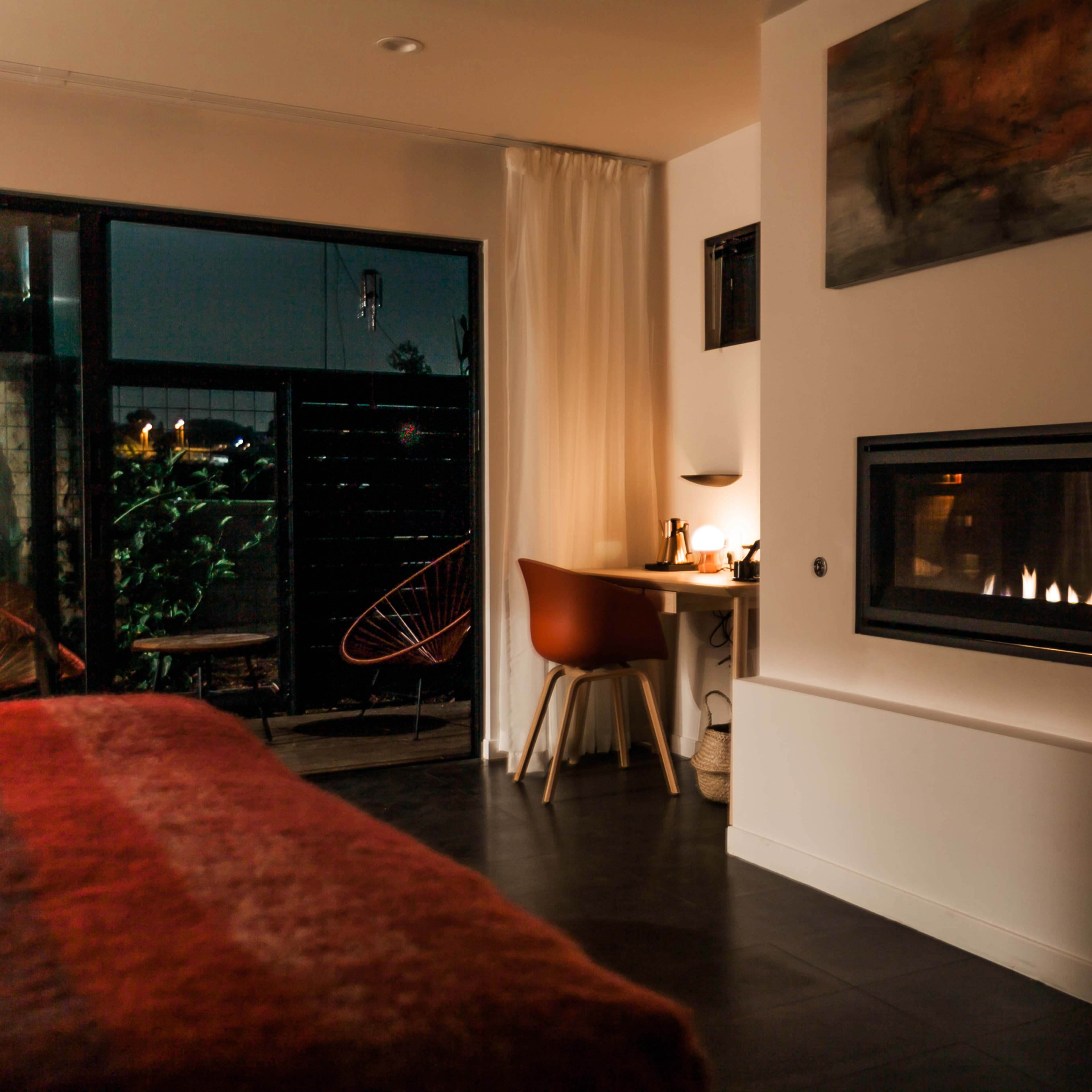 A modern bedroom with a cozy bed, a desk with a chair, and a fireplace, illuminated by soft light, alongside a glass door leading to a balcony.