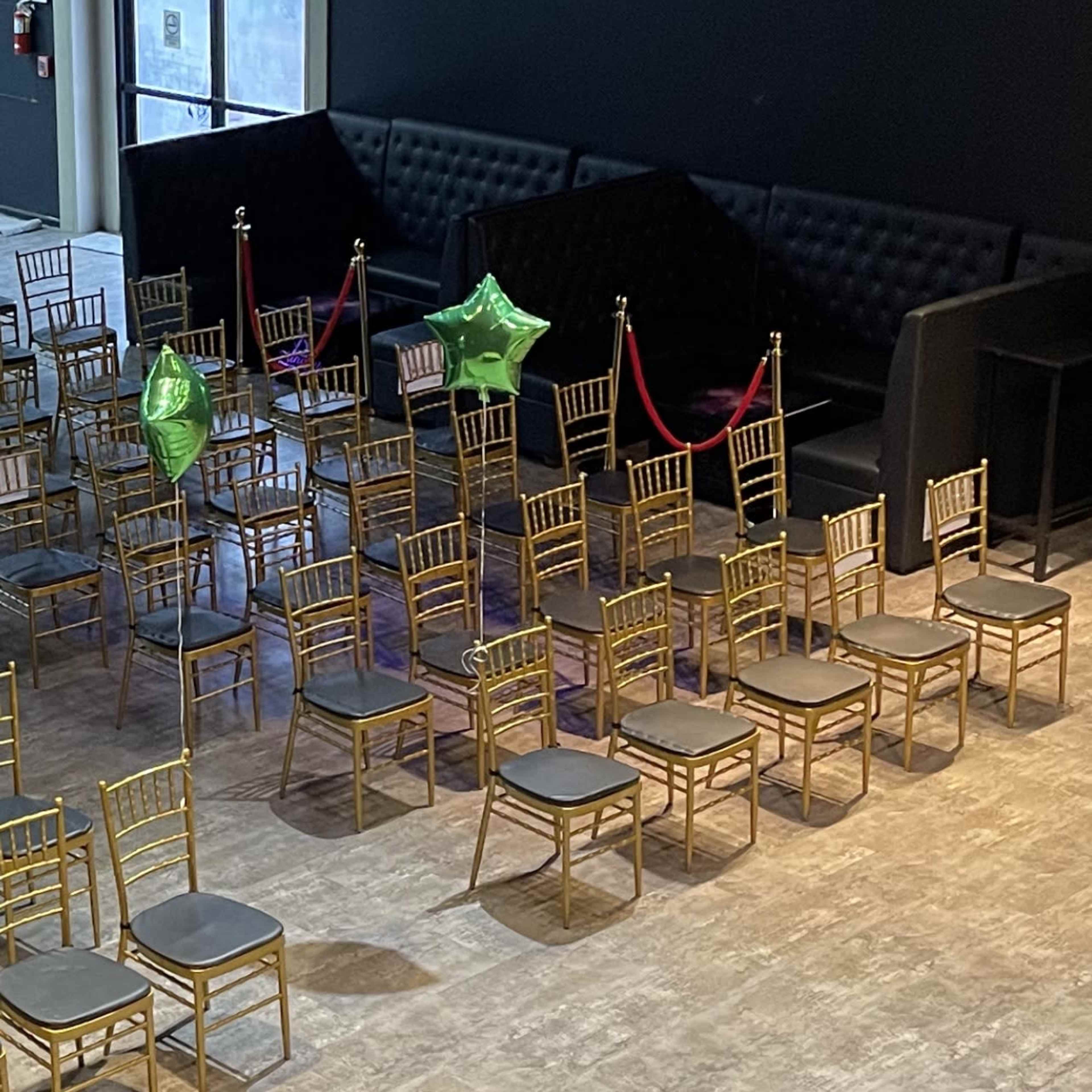 A spacious event venue with rows of empty gold chairs arranged neatly on a wooden floor, complemented by green balloons and a black backdrop.