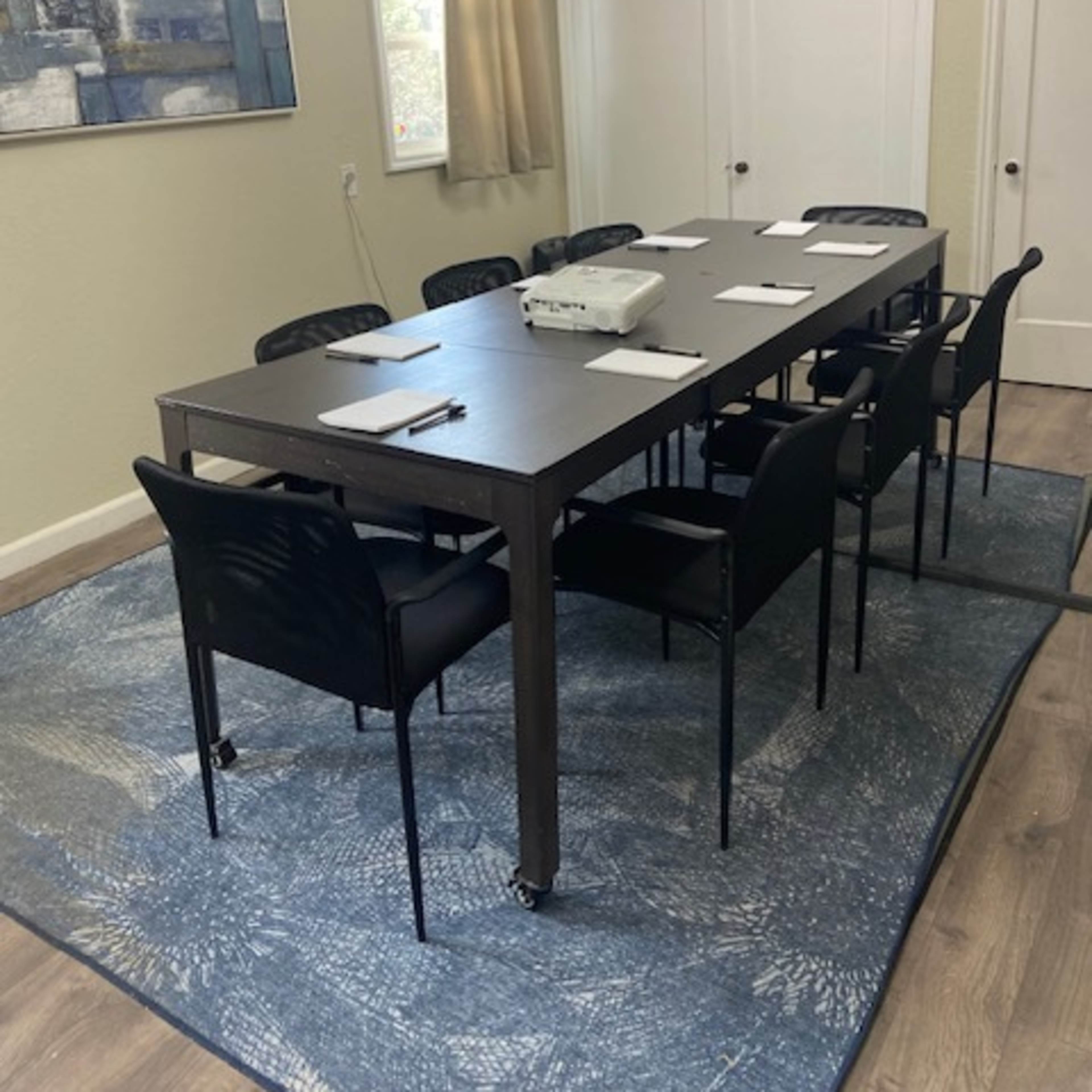 A rectangular conference table with eight black chairs is arranged on a blue rug in a well-lit room with two doors and a wall-mounted projector.