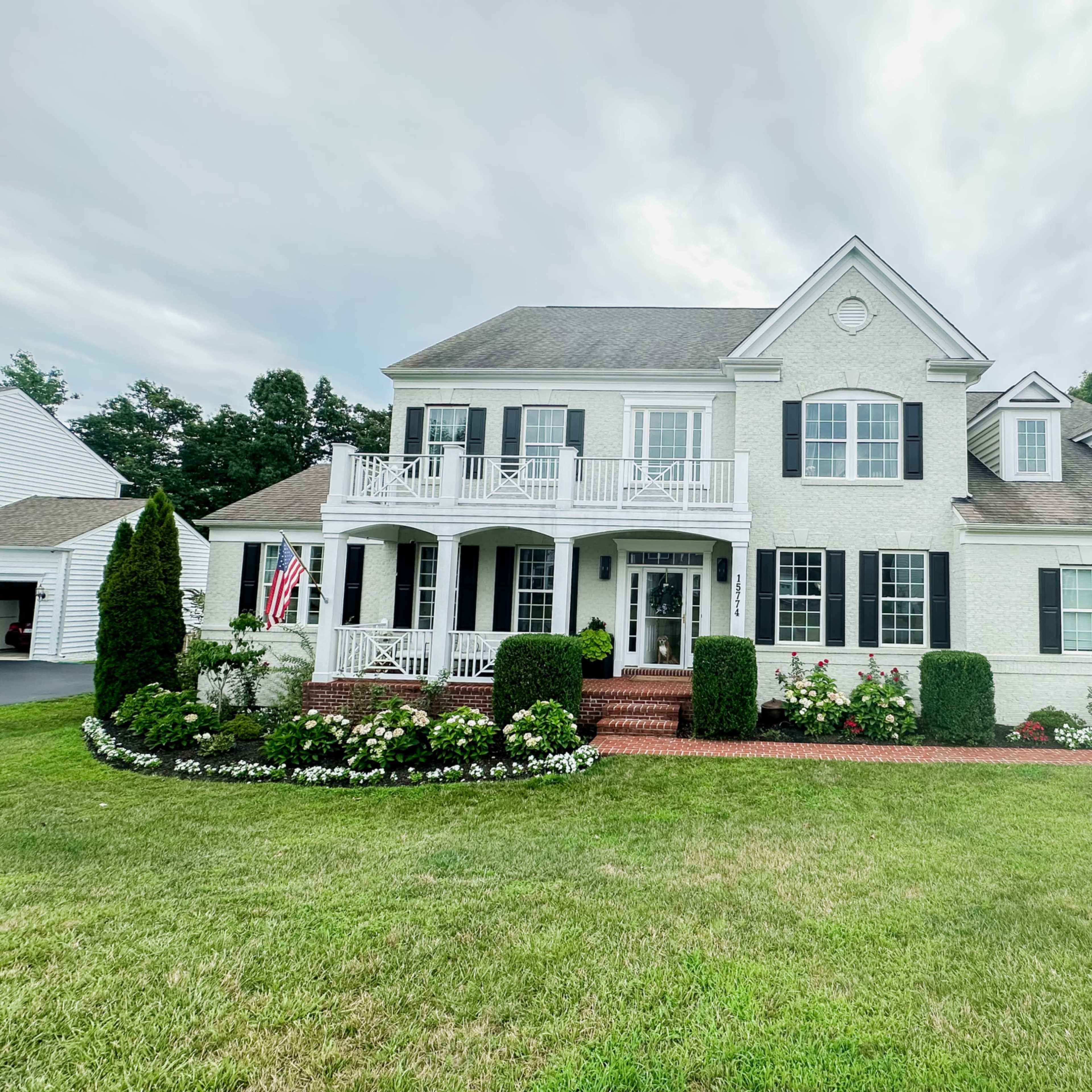 A two-story white house with black shutters, a brick walkway, and a landscaped front yard featuring flowers and a flagpole.