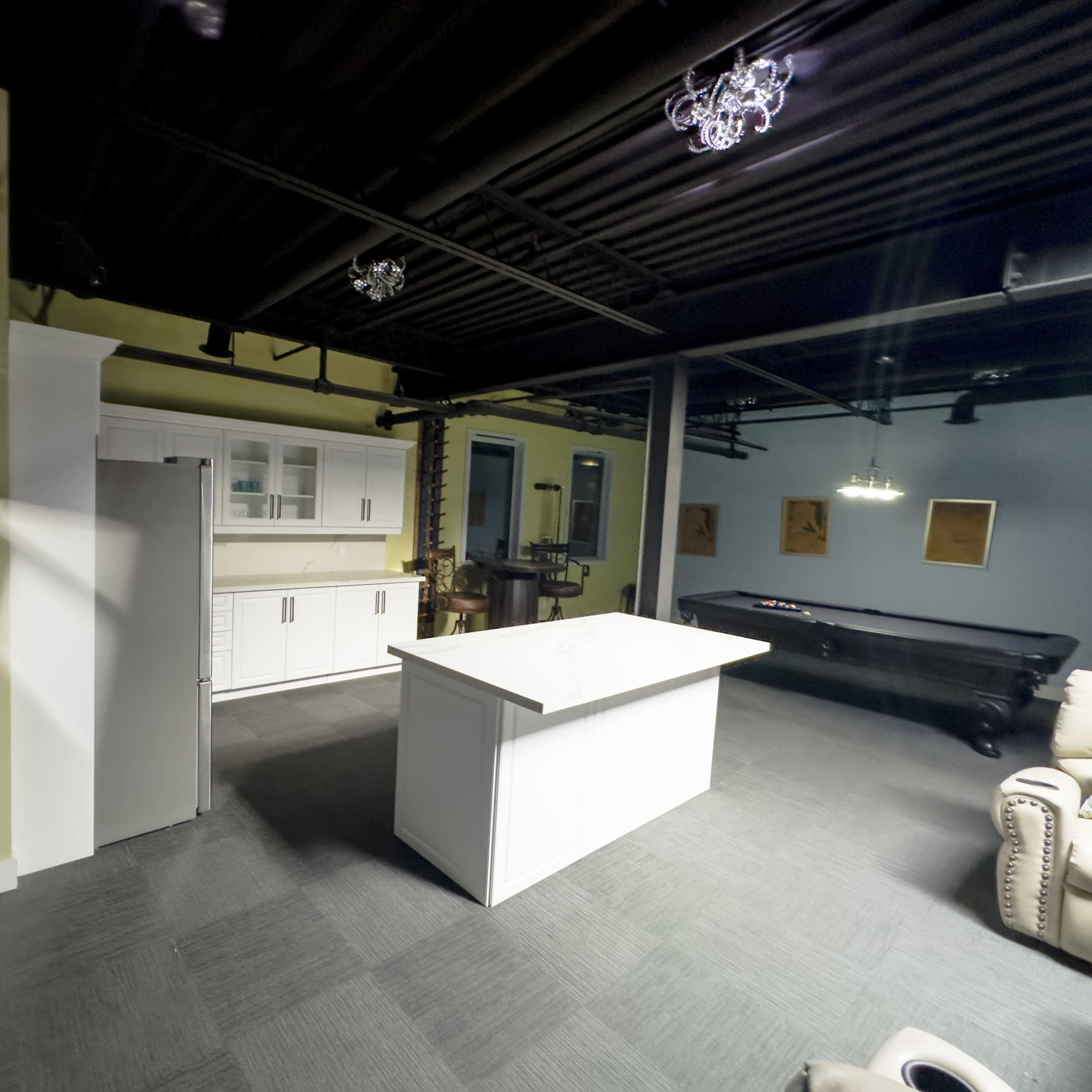 A modern kitchen area featuring white cabinetry and a central island, with a refrigerator on the left and a seating area visible in the background.
