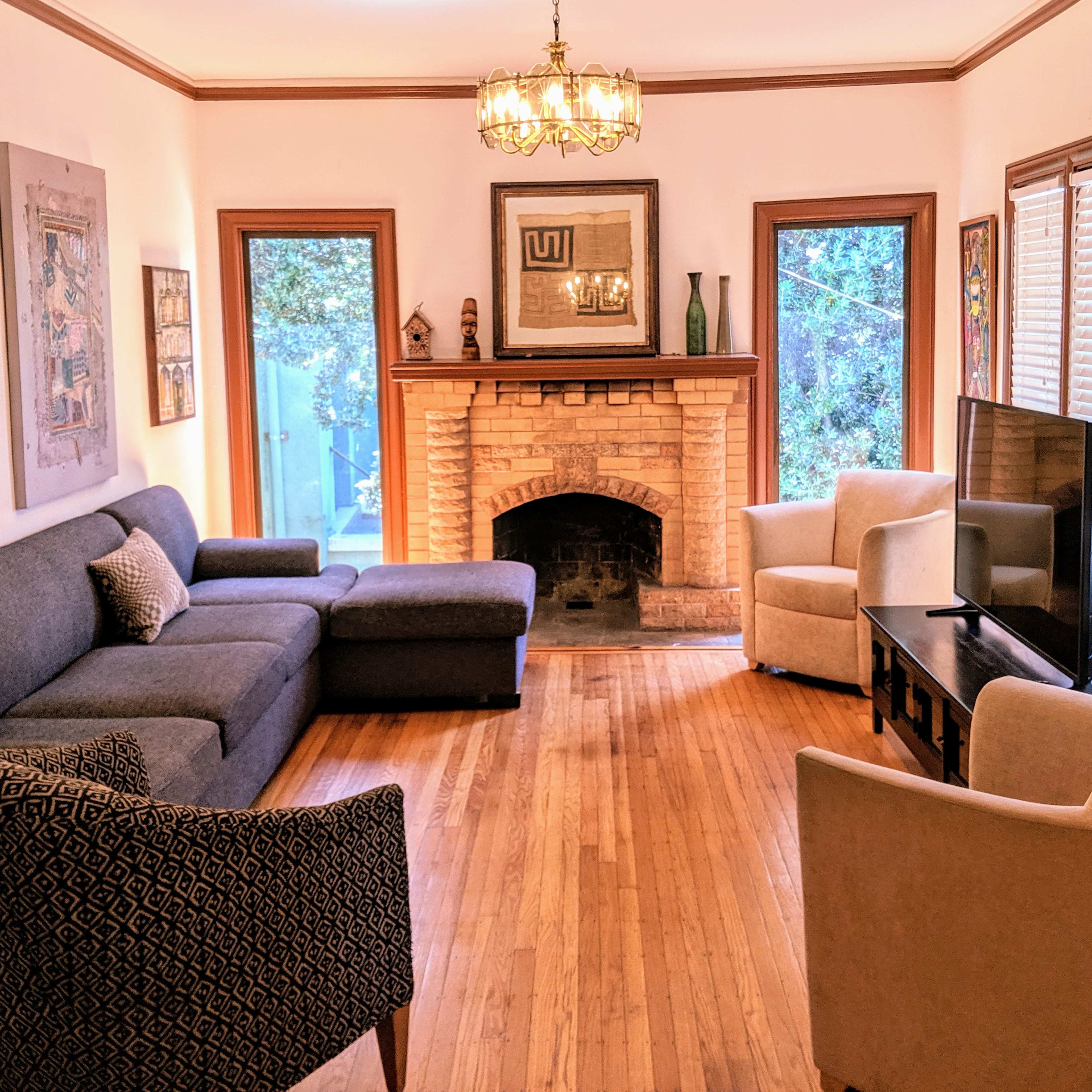 A living room features a blue sectional sofa, two cream-colored armchairs, a coffee table, and a television near a brick fireplace, with natural light coming through the windows.
