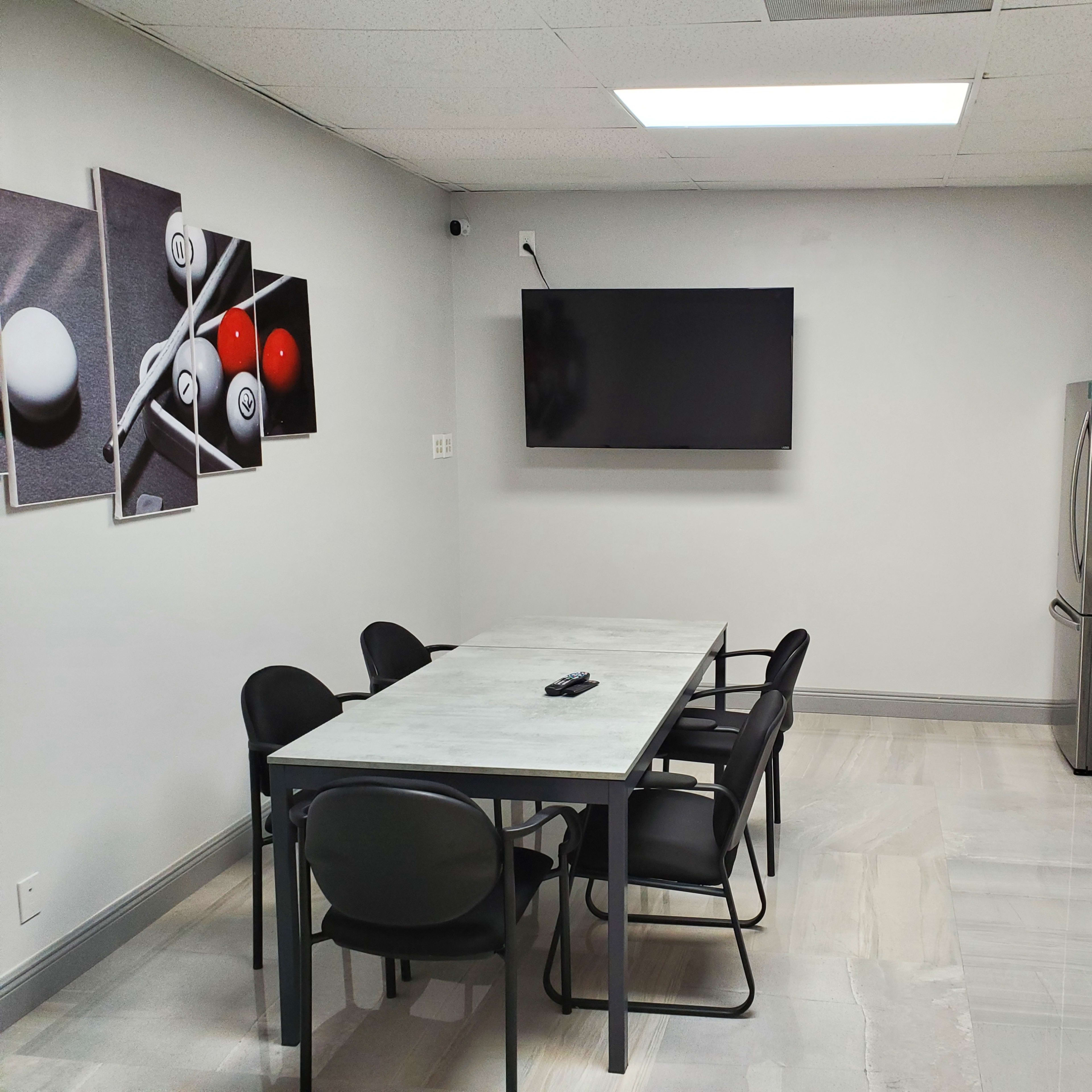 A modern break room with a table and chairs, a kitchenette with stainless steel appliances, and a large TV mounted on the wall.