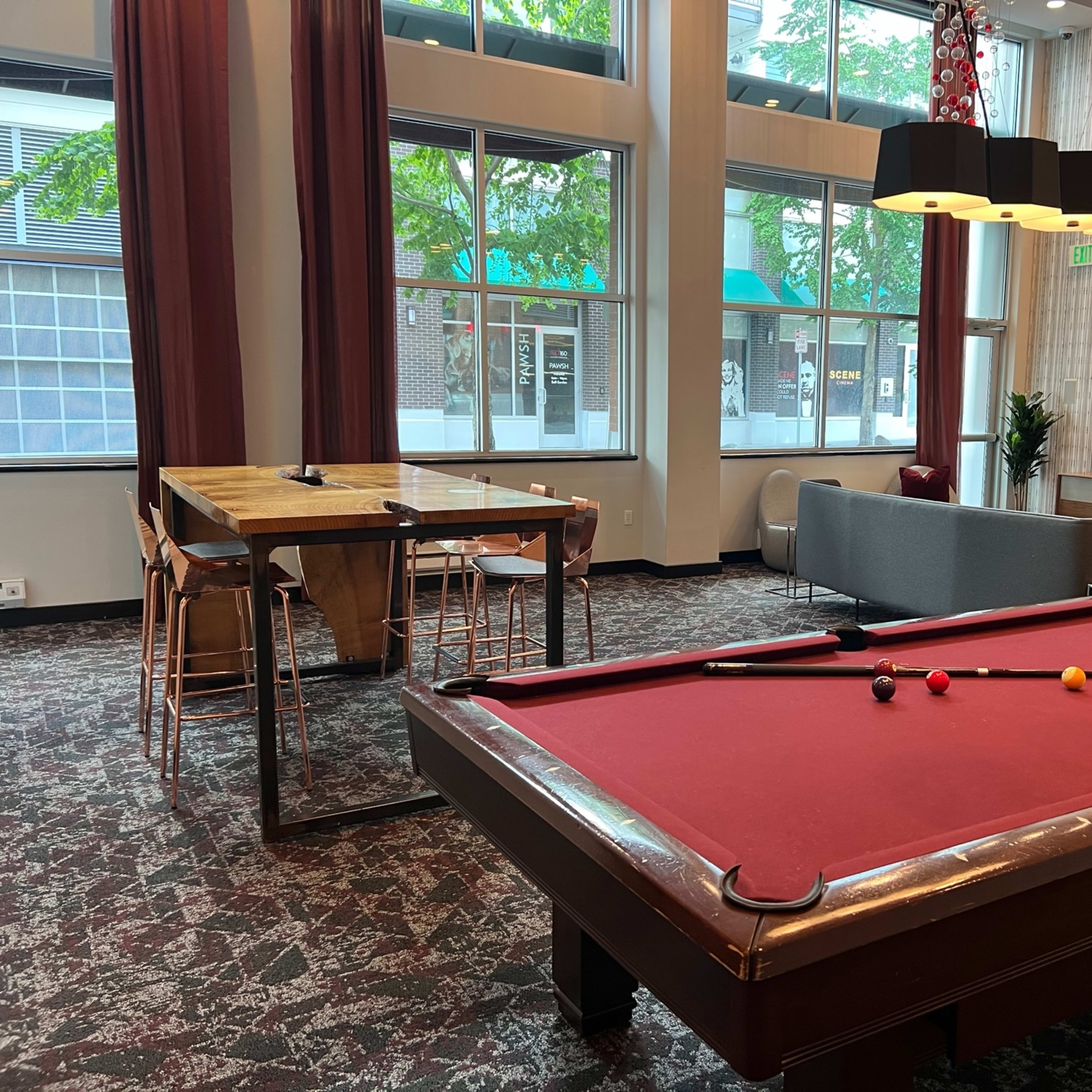 A recreational lounge area featuring a pool table in the foreground, with a dining table and chairs, a sofa, and a television in a well-lit space with large windows.