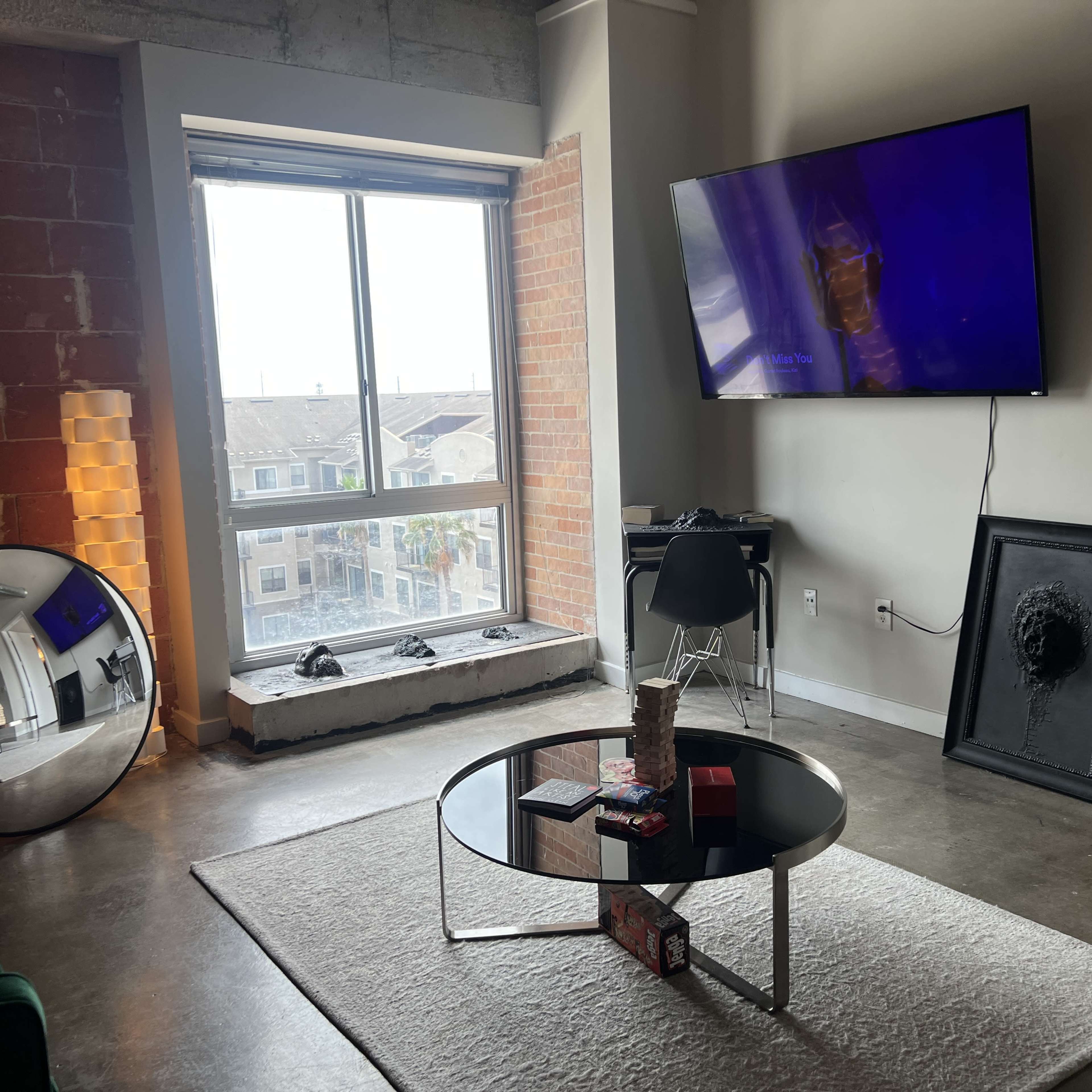 A modern living room with a large window, a round coffee table, a TV mounted on the wall, and a mirror reflecting the space.