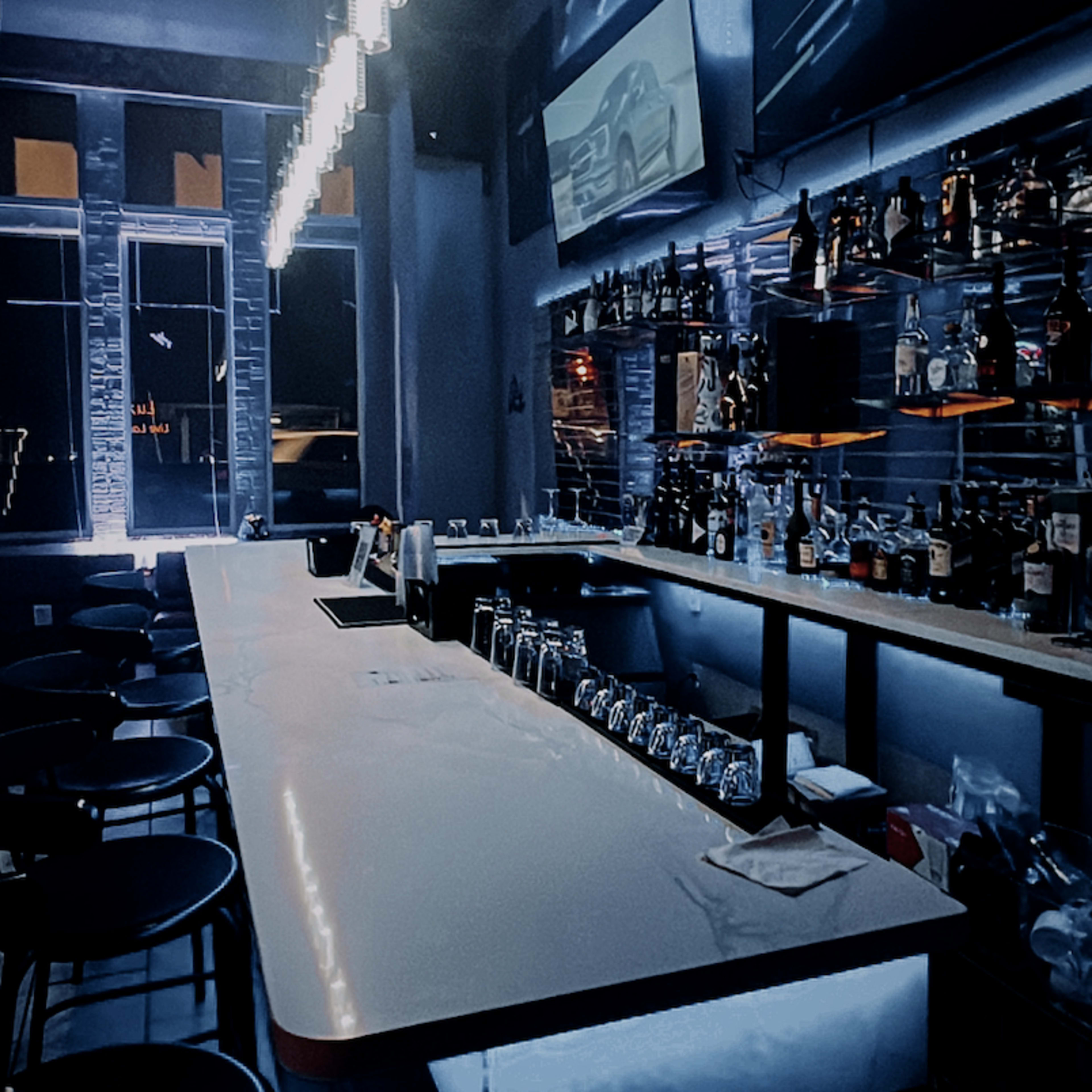 A modern bar with a long countertop, illuminated shelves displaying bottles, and ambient lighting.
