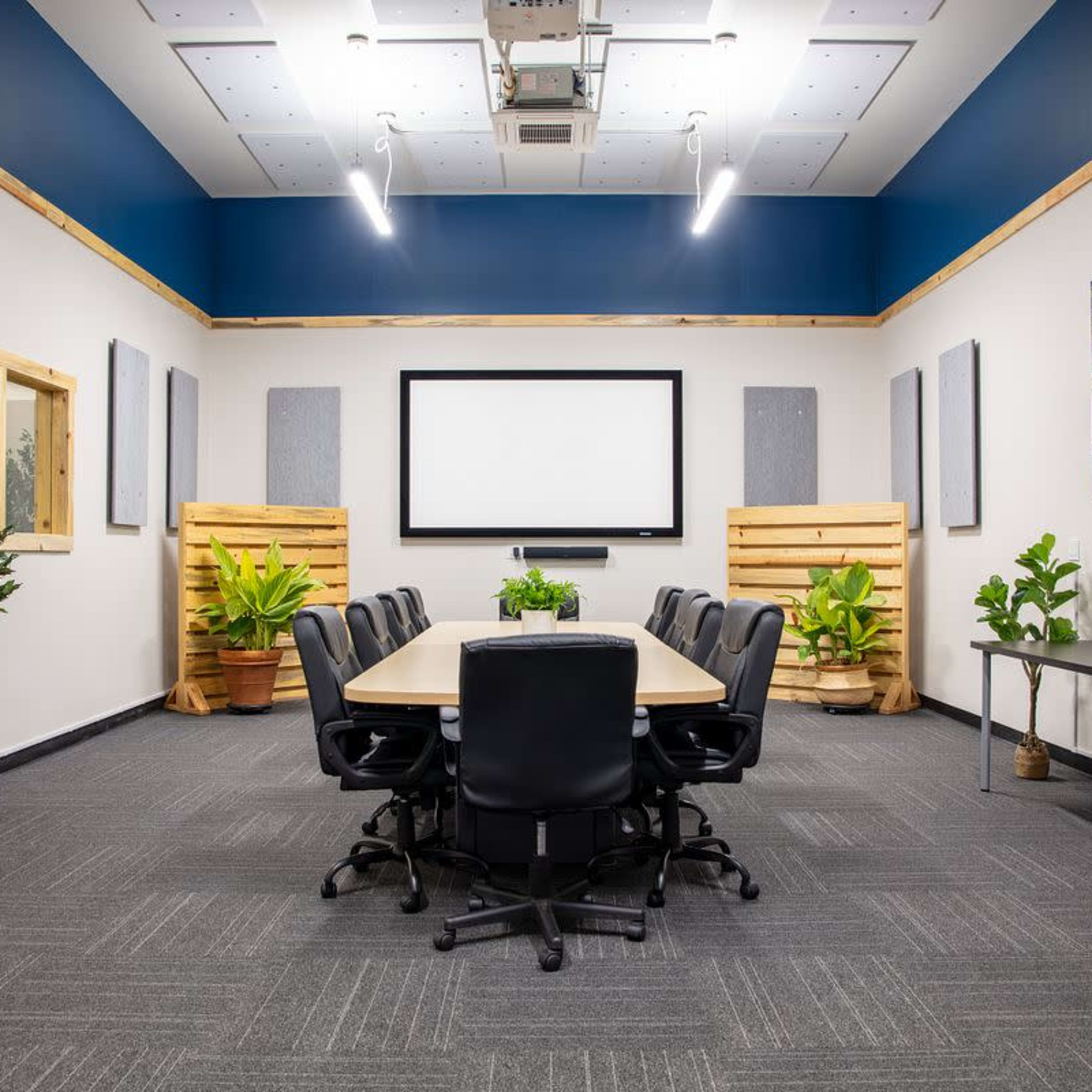 A conference room is equipped with a rectangular table and black chairs, flanked by plants and acoustic panels, with a large screen at the front and artwork on the walls.
