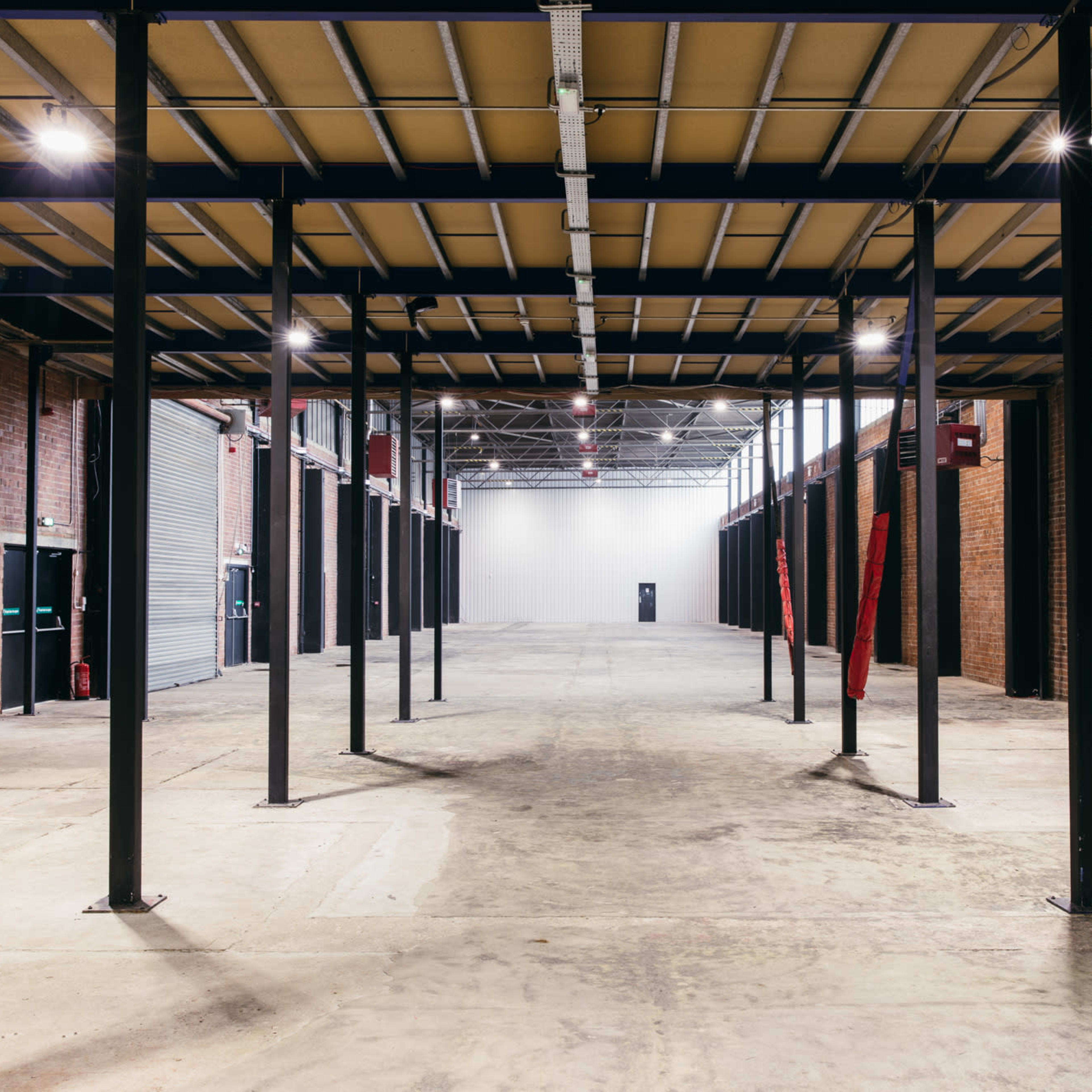 A spacious, empty warehouse features exposed brick walls and steel support beams, with minimal lighting and a concrete floor.