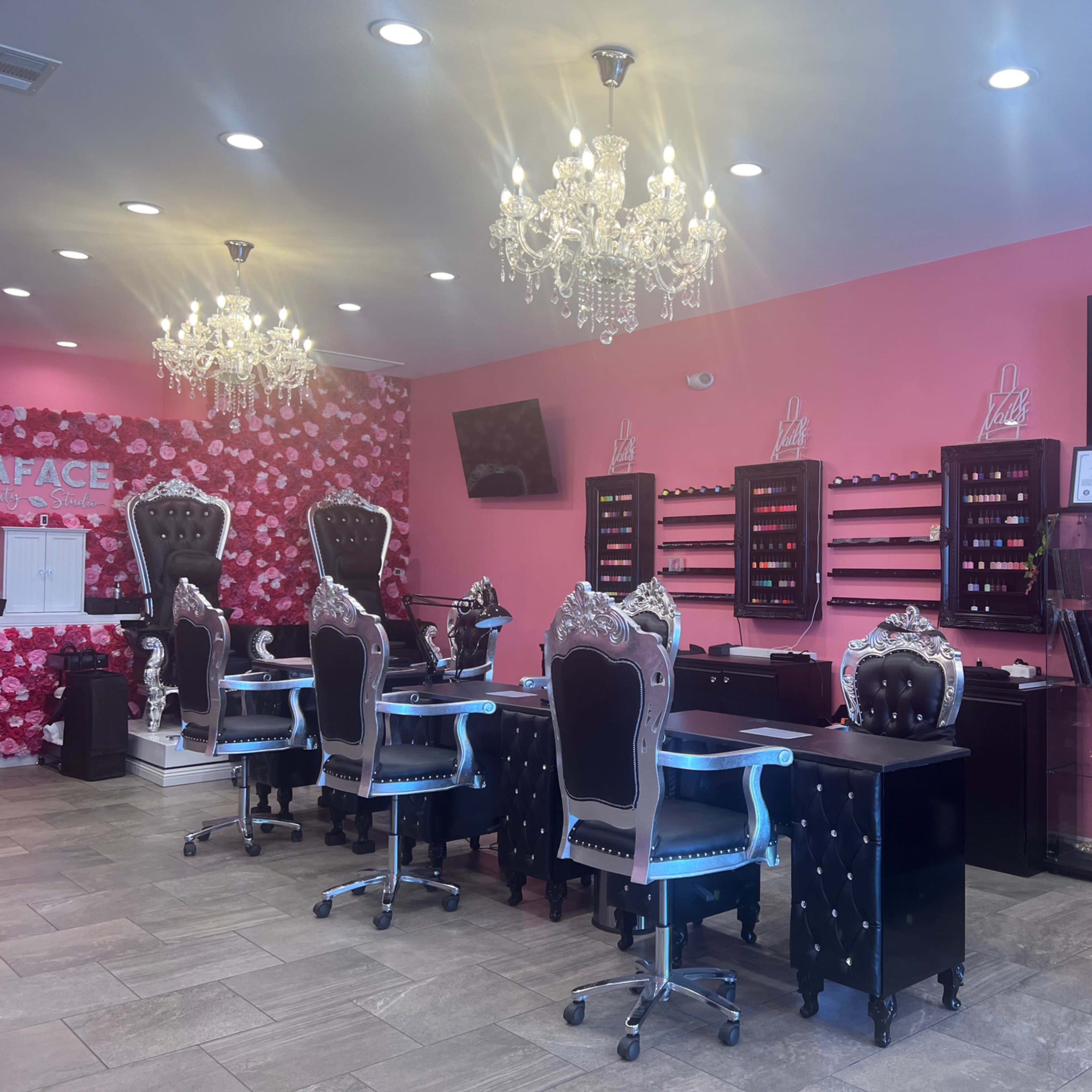 The interior of a nail salon featuring black and silver chairs, a pink wall adorned with floral decorations, and chandeliers hanging from the ceiling.