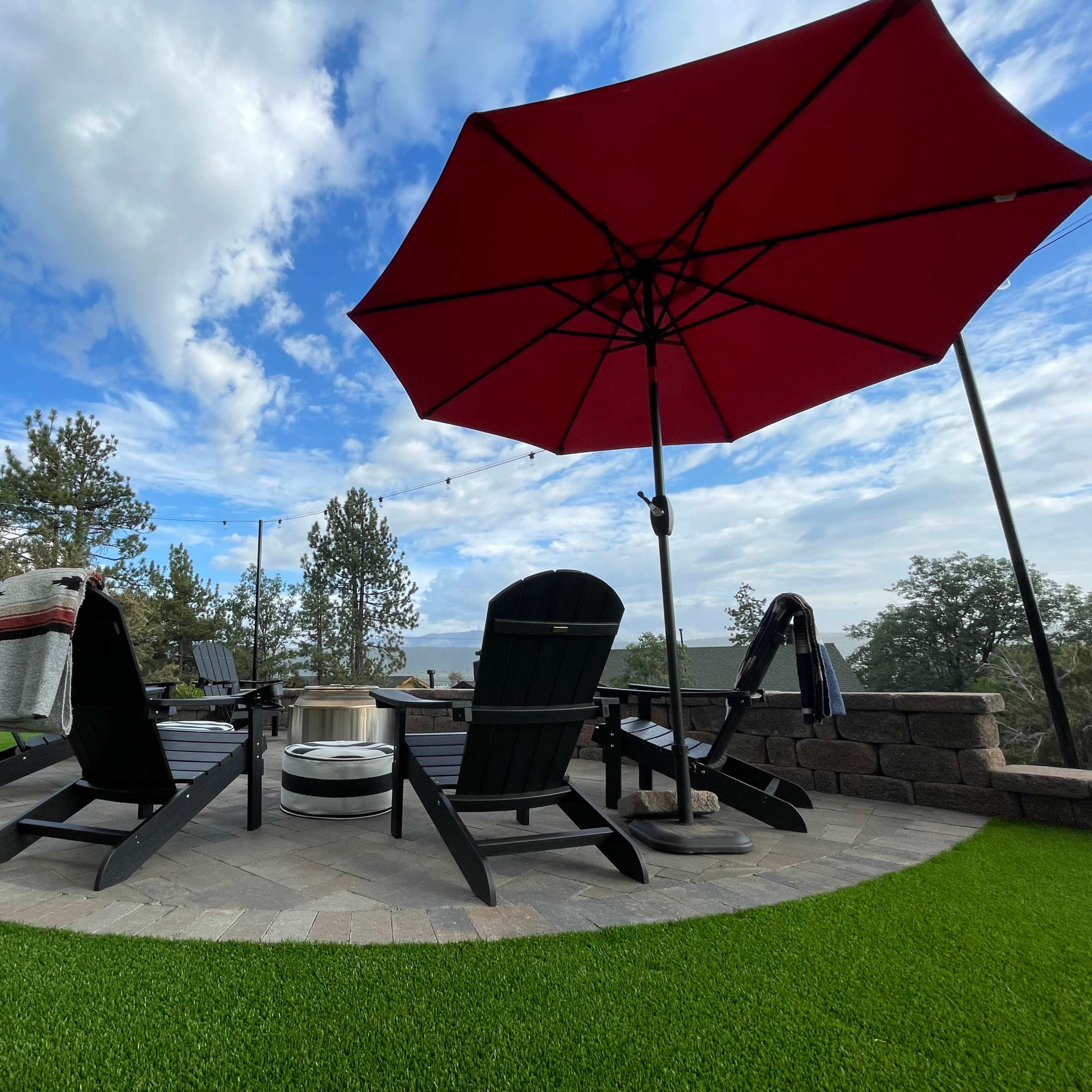 A circular stone patio features black Adirondack chairs, a fire pit, and a red umbrella on a lawn of artificial grass.