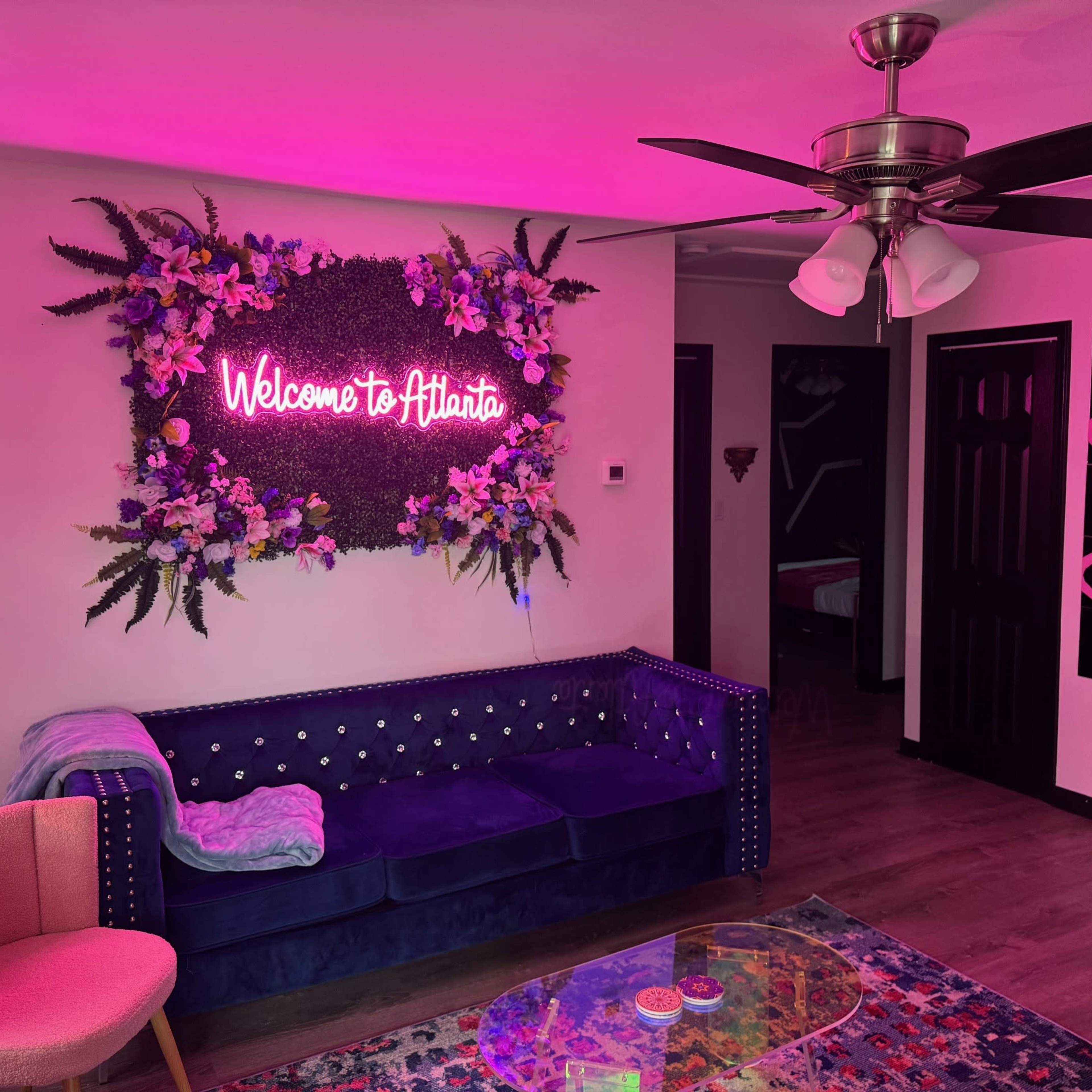 A cozy interior space with a blue velvet couch, a pink neon sign that says "Welcome to Atlanta," and colorful decor elements including a patterned rug and modern lighting.