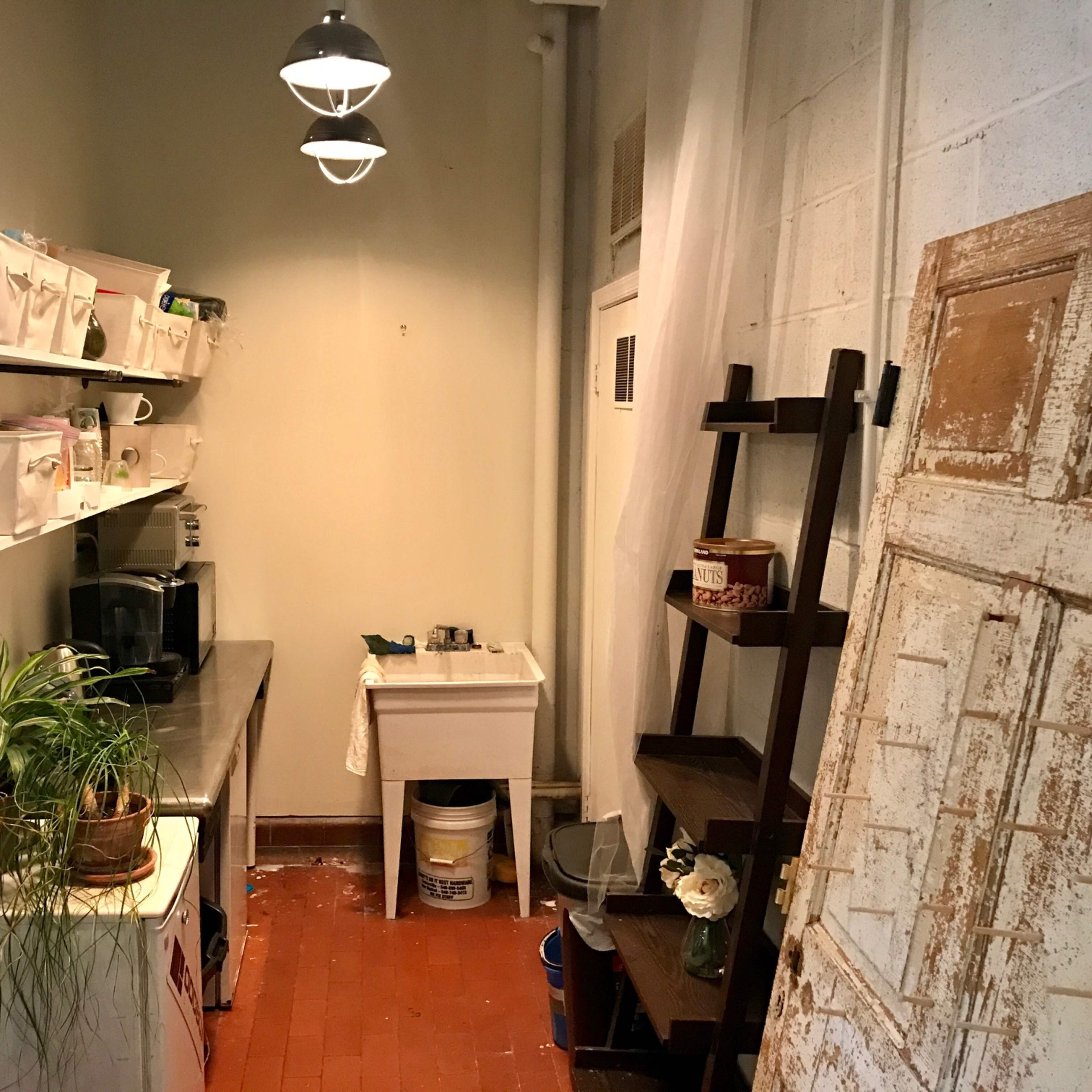A small kitchen area with a stainless steel countertop, shelves containing storage boxes, a sink, and a wooden door leaning against the wall.