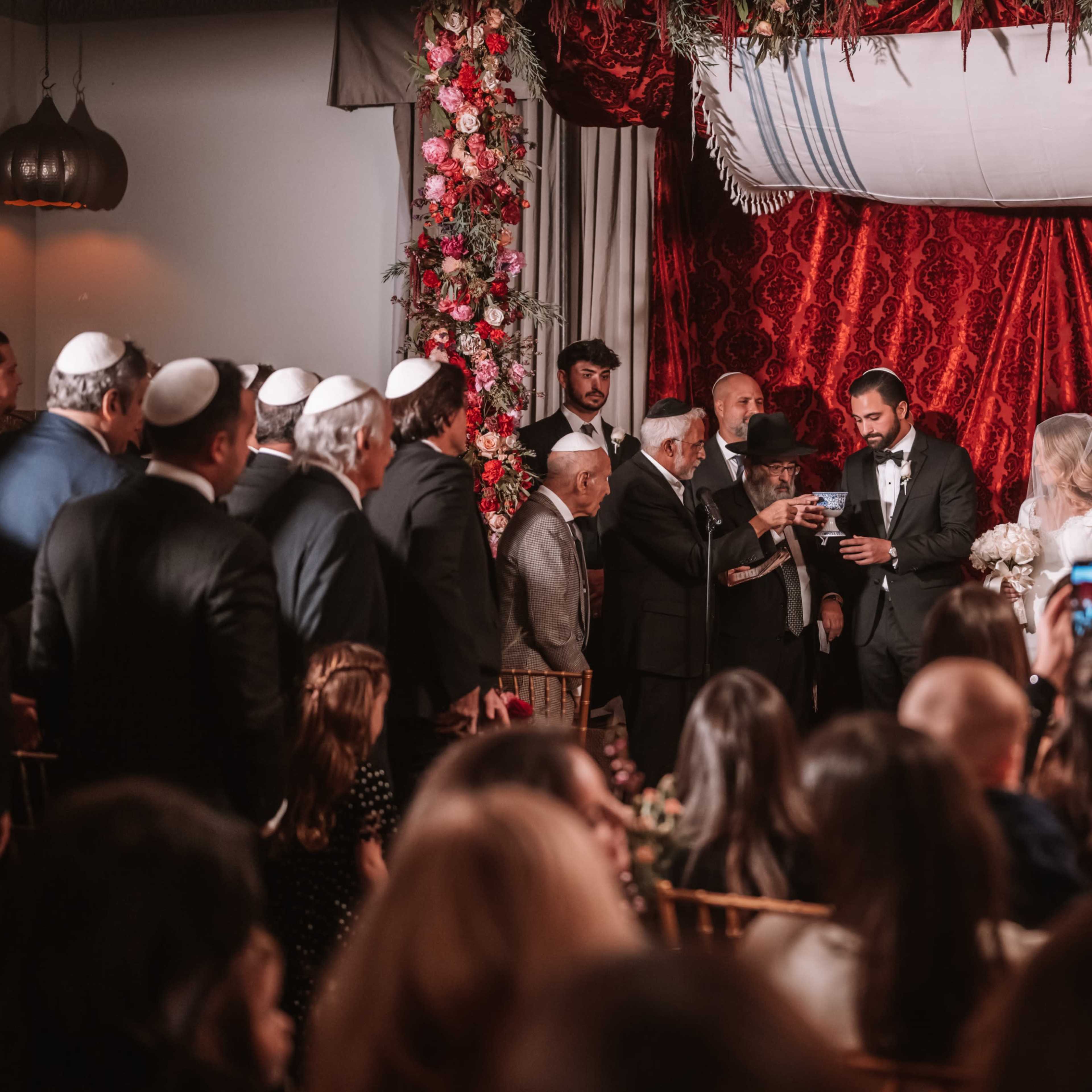 A wedding ceremony is taking place under a decorated canopy, with guests seated in rows while the couple stands before officiants.