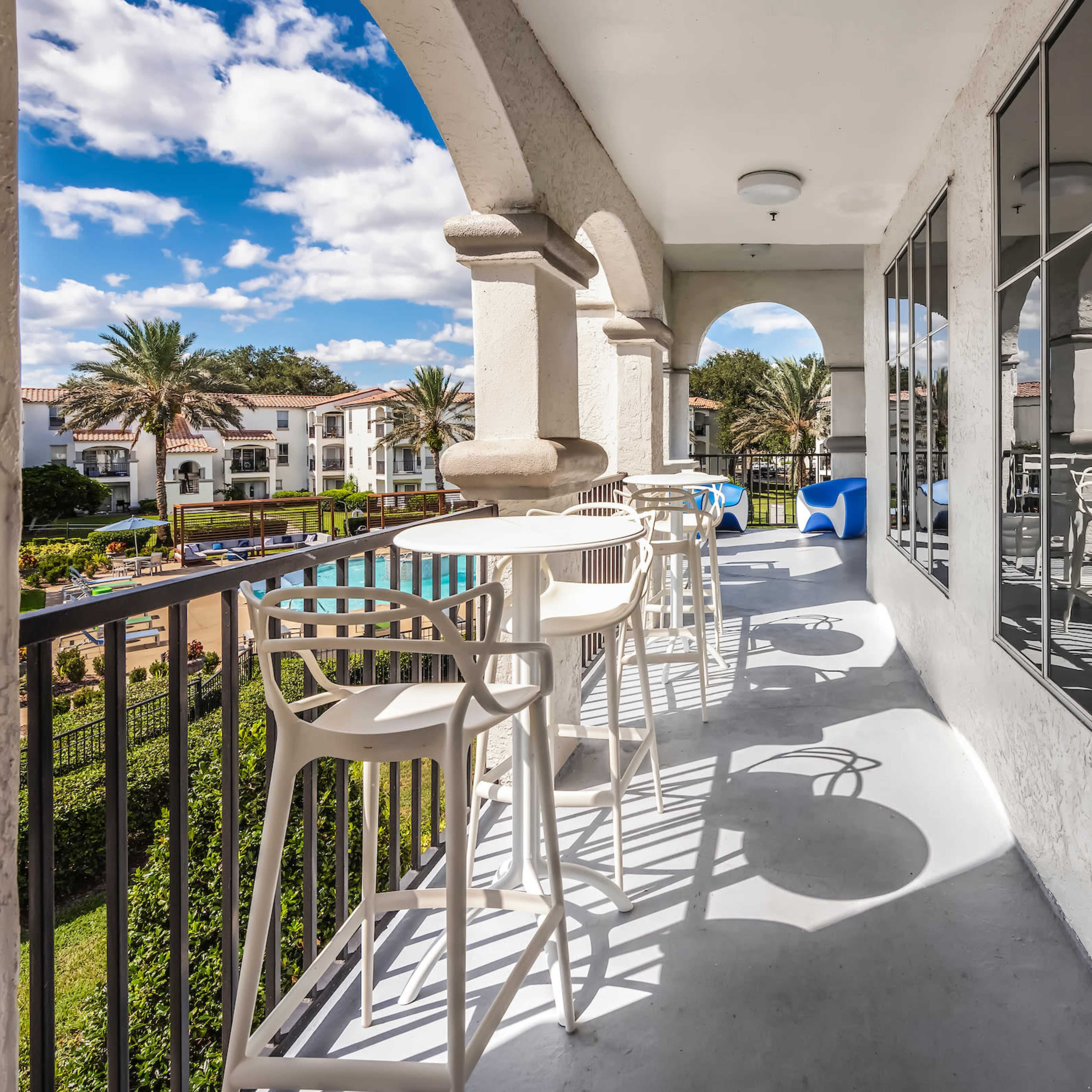 A balcony with modern bar stools overlooking a pool area surrounded by palm trees and several buildings.