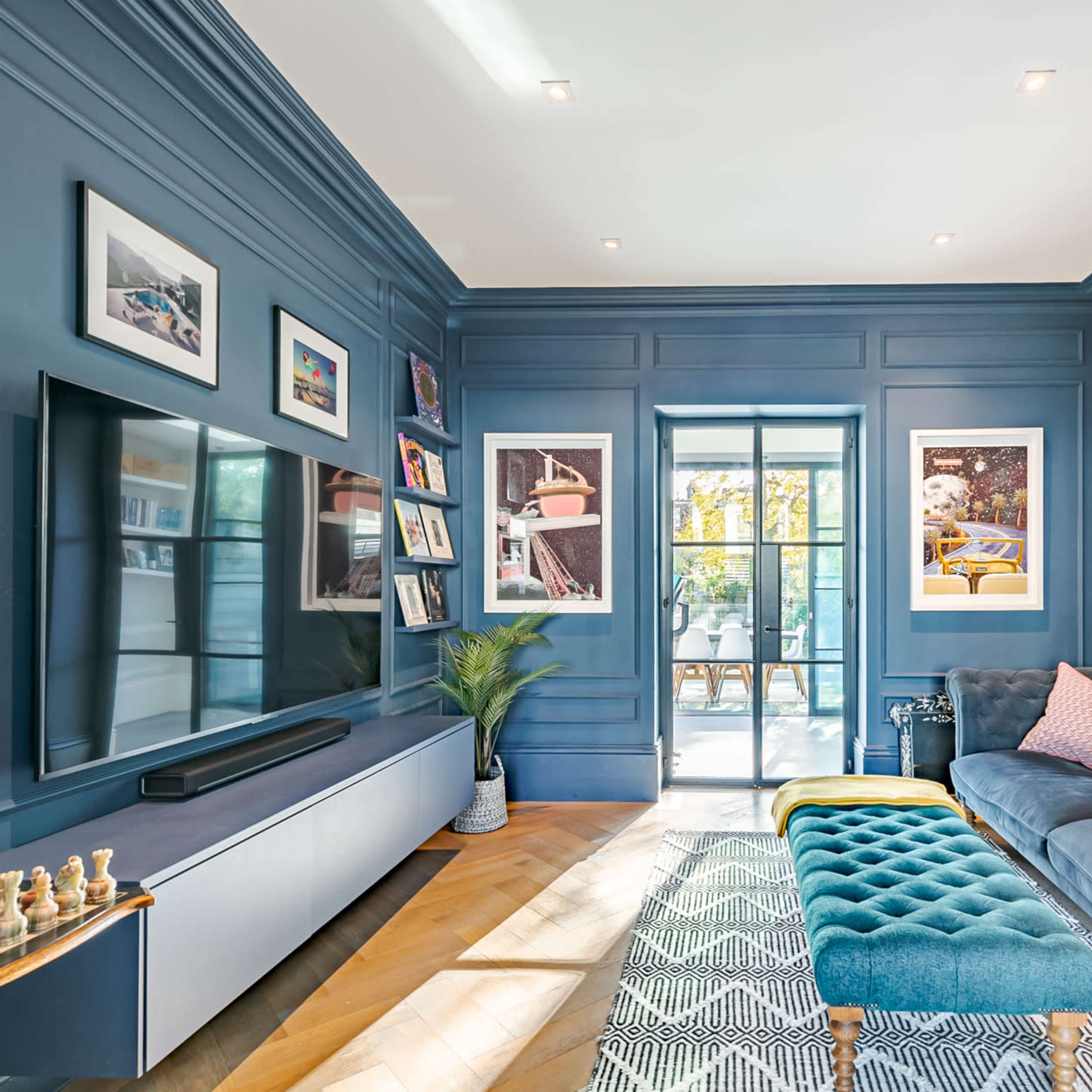 A modern living room with blue walls, a large flat-screen TV, a tufted sofa, and artwork hanging on the walls, leading to a door that opens to an outdoor space.
