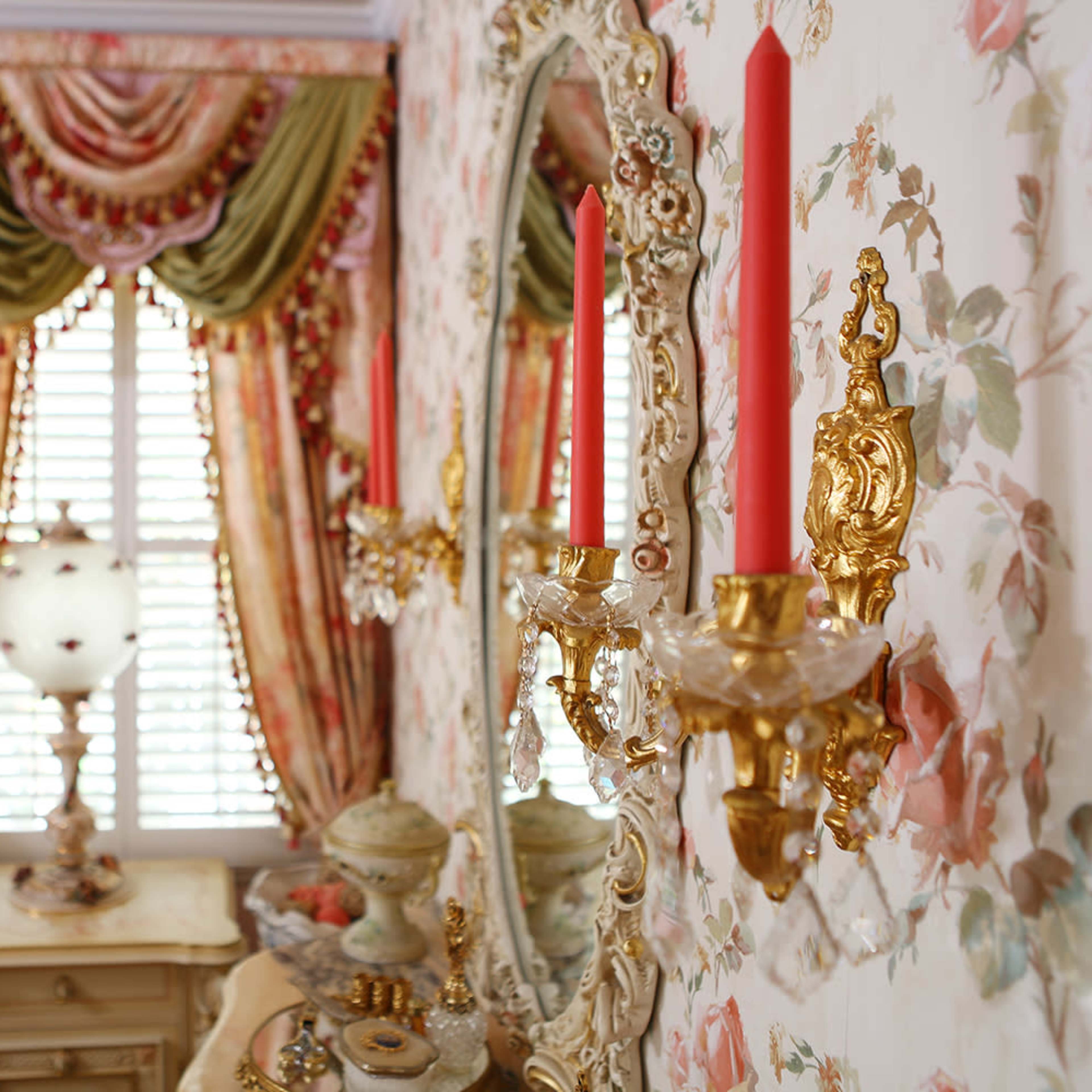 A elegantly decorated bedroom features floral wallpaper, ornate mirrors, and candle sconces flanking a window covered with draped curtains.